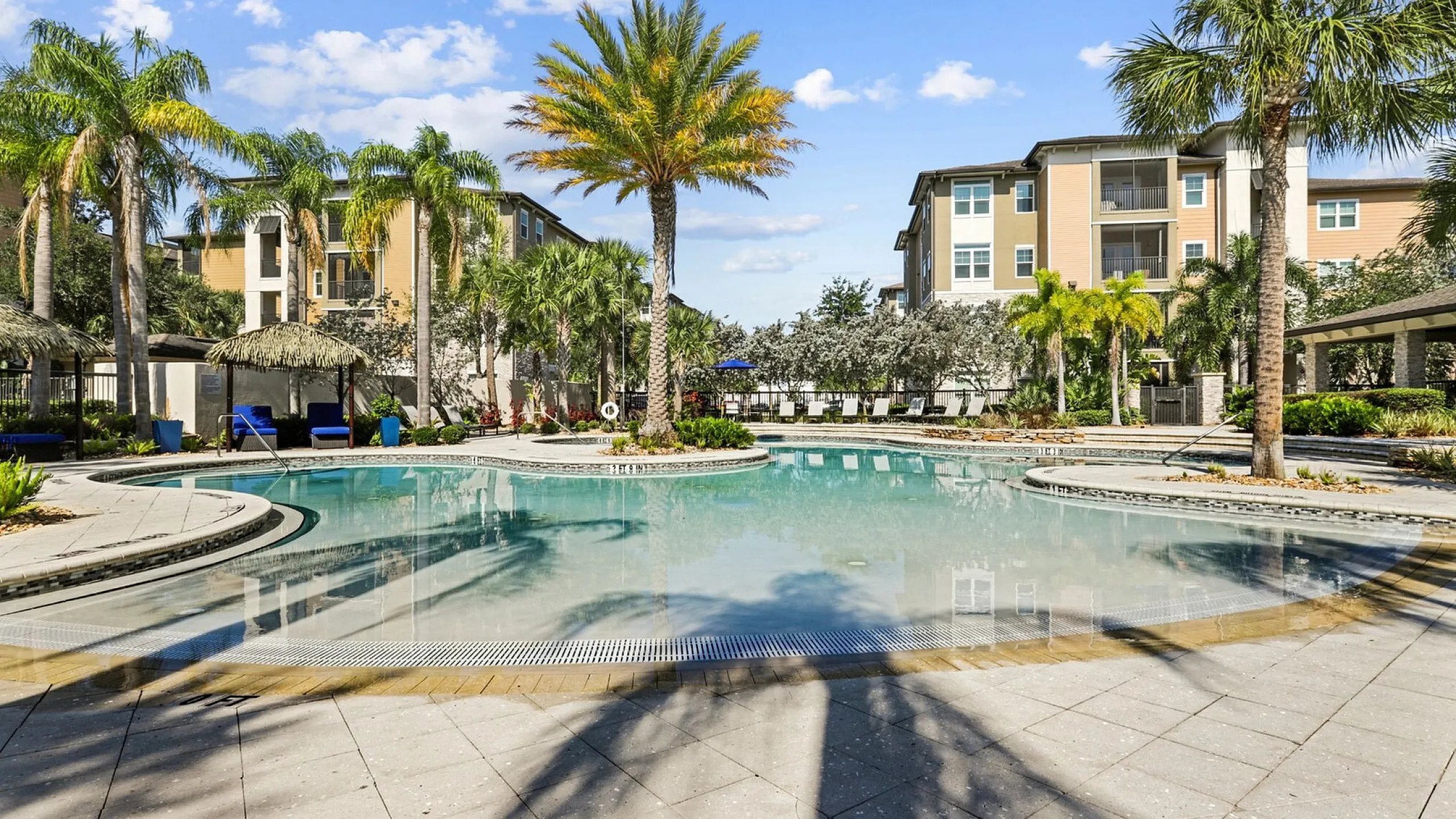 Luxurious apartment complex with stunning resort-style pool, surrounded by lush palm trees and elegant buildings under a clear blue sky.