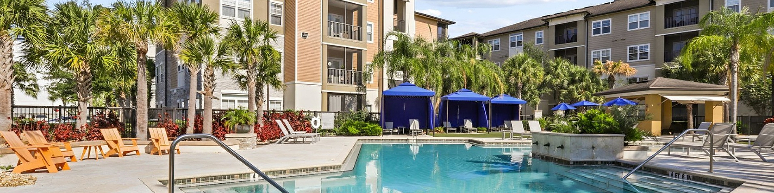 Luxurious apartment complex featuring a sparkling pool with palm trees, comfortable lounge chairs, and elegant blue cabanas, perfect for relaxation and enjoyment.