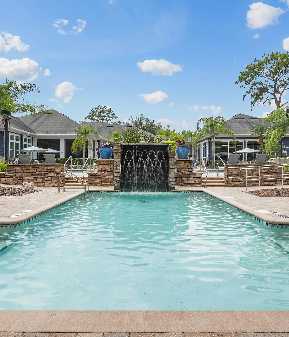 Luxurious apartment complex pool area featuring a sparkling swimming pool with waterfall, surrounded by lush greenery and elegant lounge chairs under a bright blue sky.