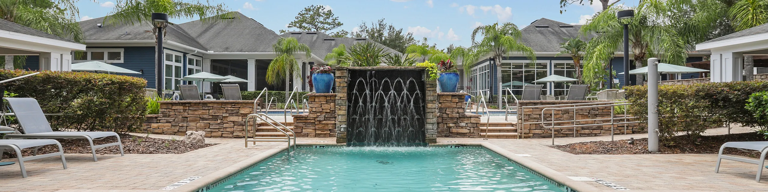 Luxurious apartment complex pool area with sparkling water, elegant waterfall feature, and surrounded by lush palm trees under a bright blue sky.