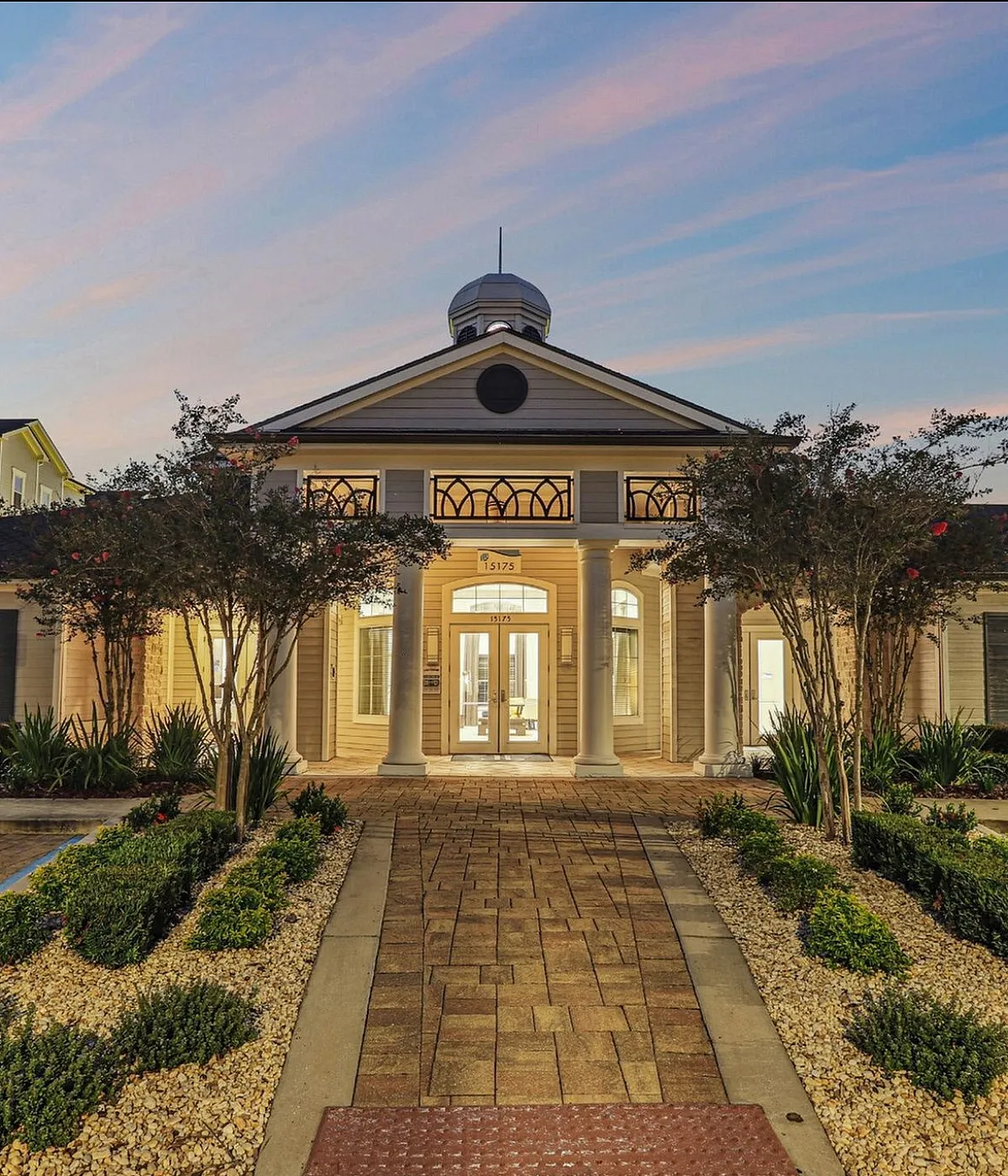Charming, well-lit entrance of a luxurious apartment complex at dusk, featuring elegant columns and manicured landscaping.