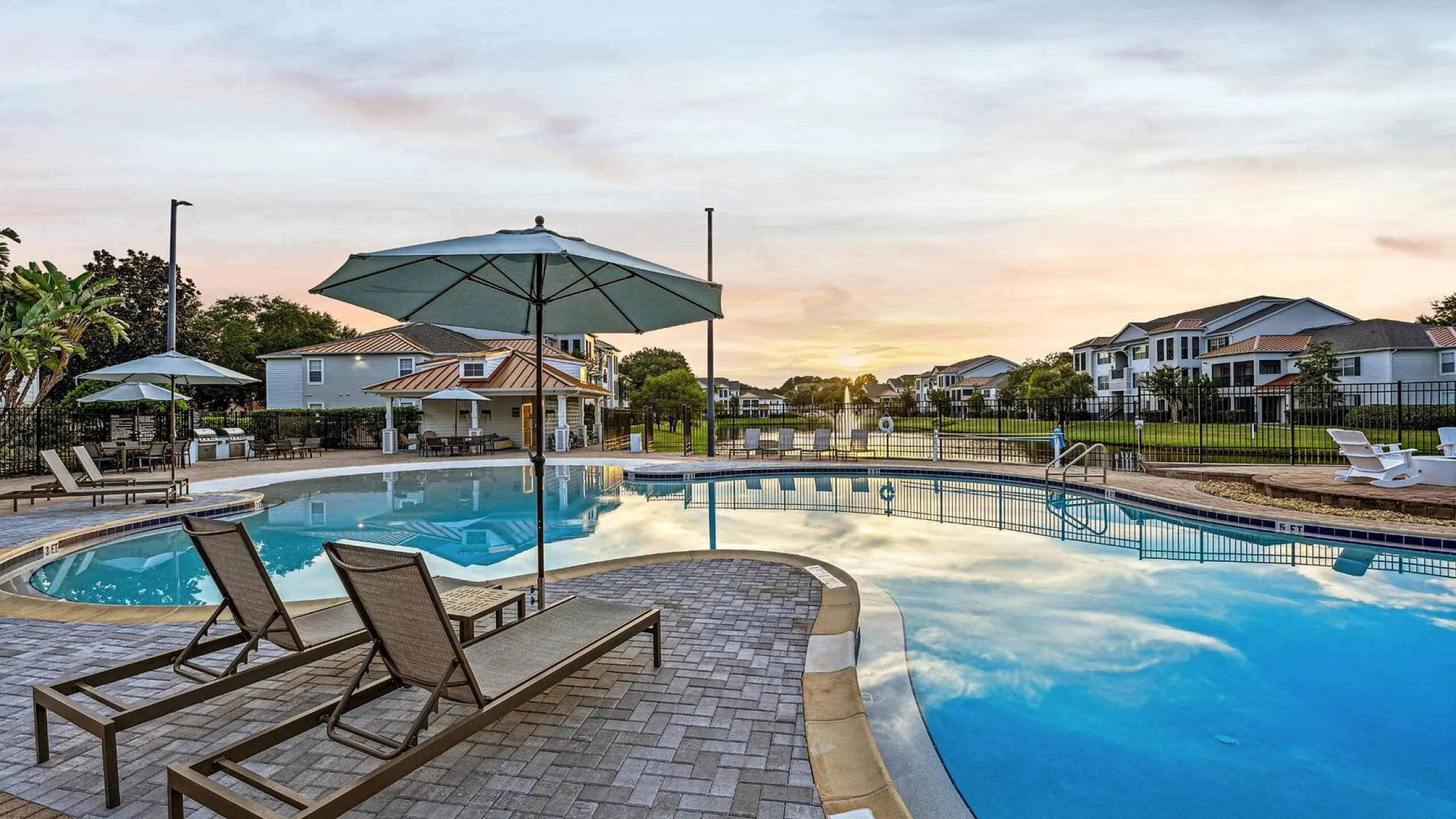 Luxurious apartment community with a stunning poolside view, featuring elegant lounge chairs and umbrellas, set against a serene sunset backdrop.