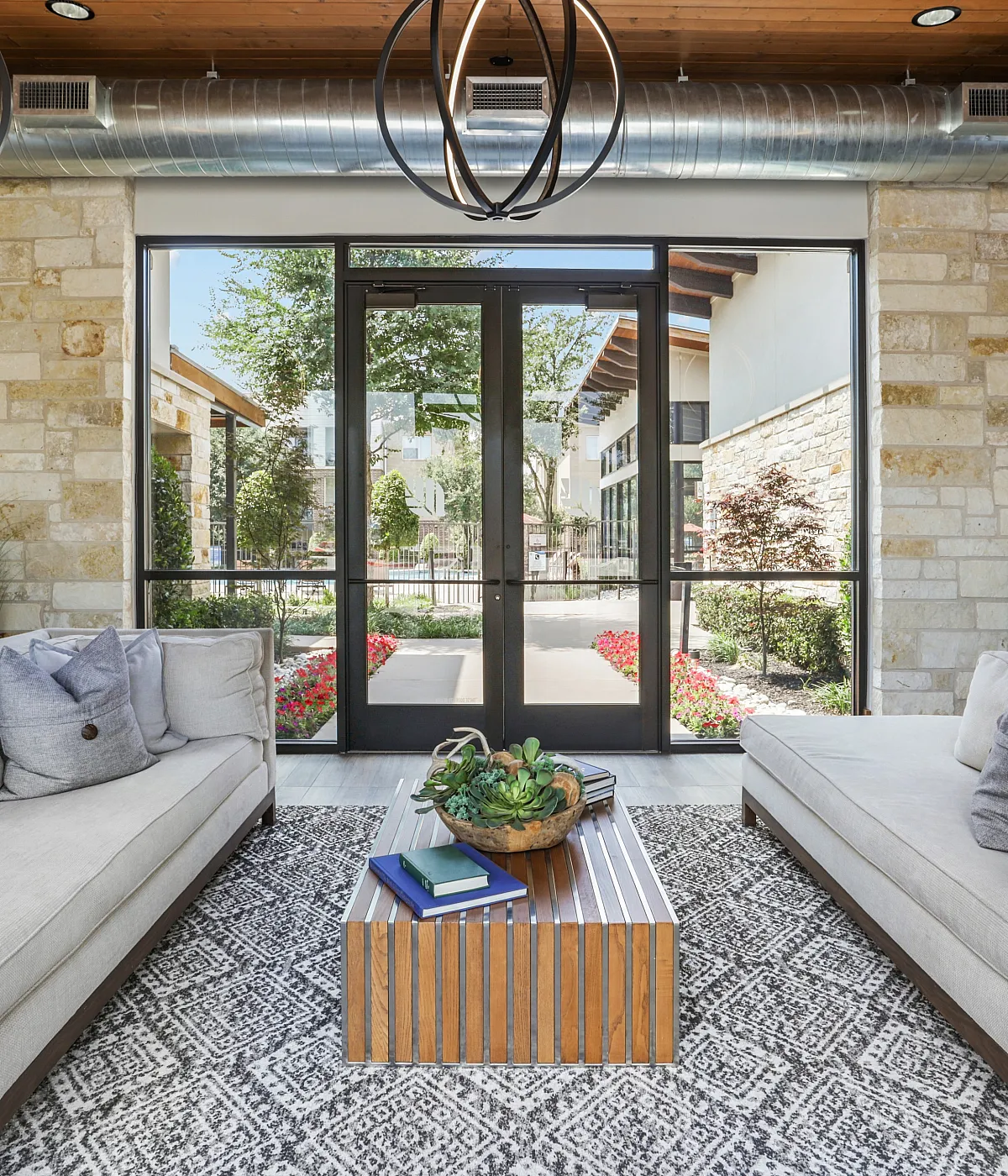 Chic lobby with modern industrial accents, featuring cozy beige sofas, a geometric patterned rug, and large windows showcasing a vibrant garden view.