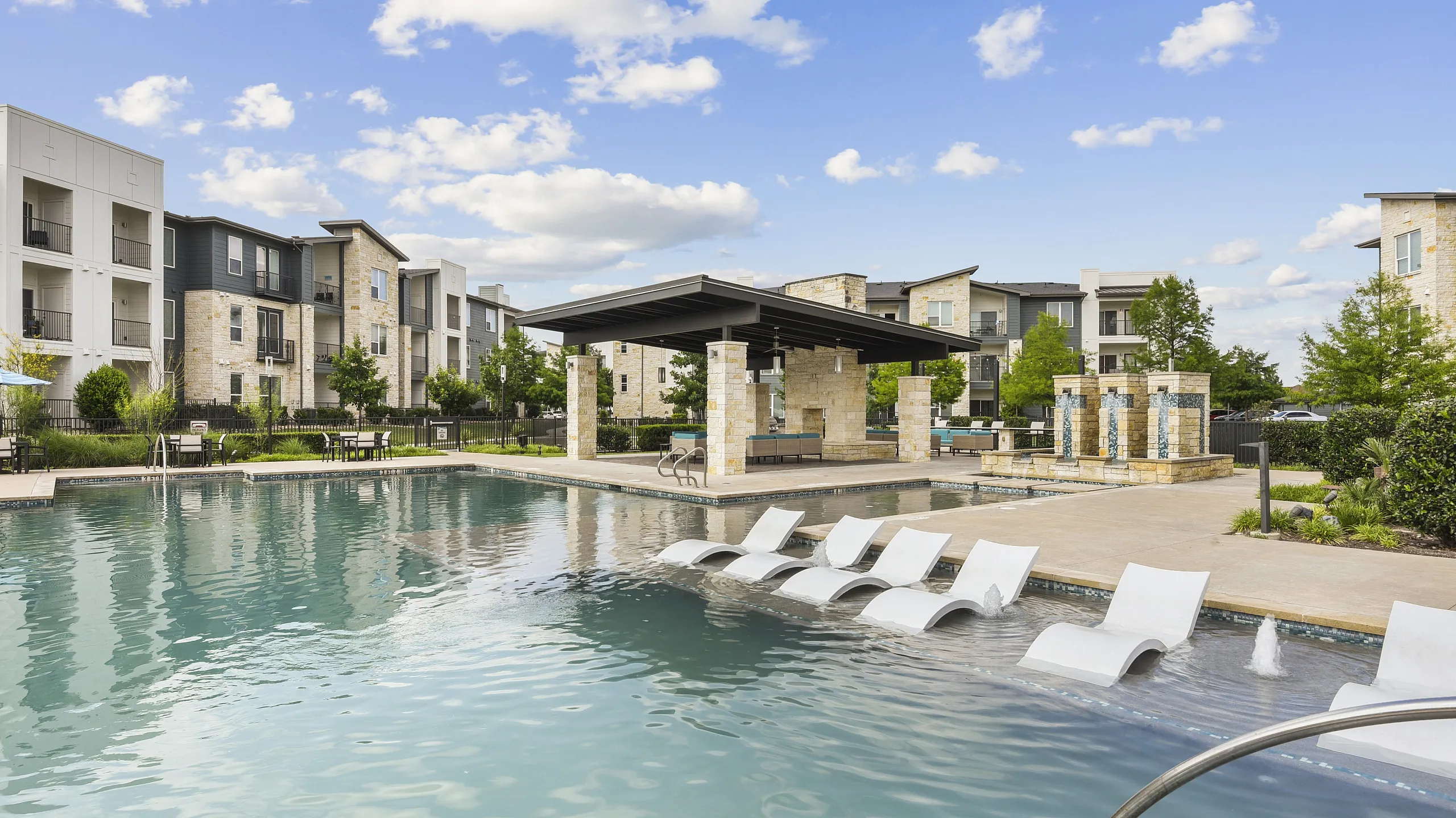 Elegant apartment complex with a luxurious poolside lounge, sunlit deck chairs, and modern architecture under a blue sky.
