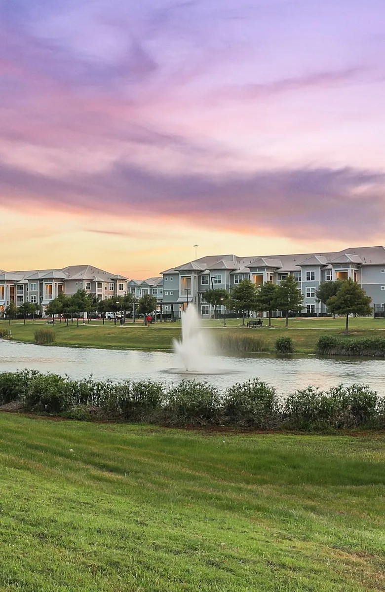 Beautiful sunset view of modern apartment complex with serene walking path and tranquil pond with fountains.