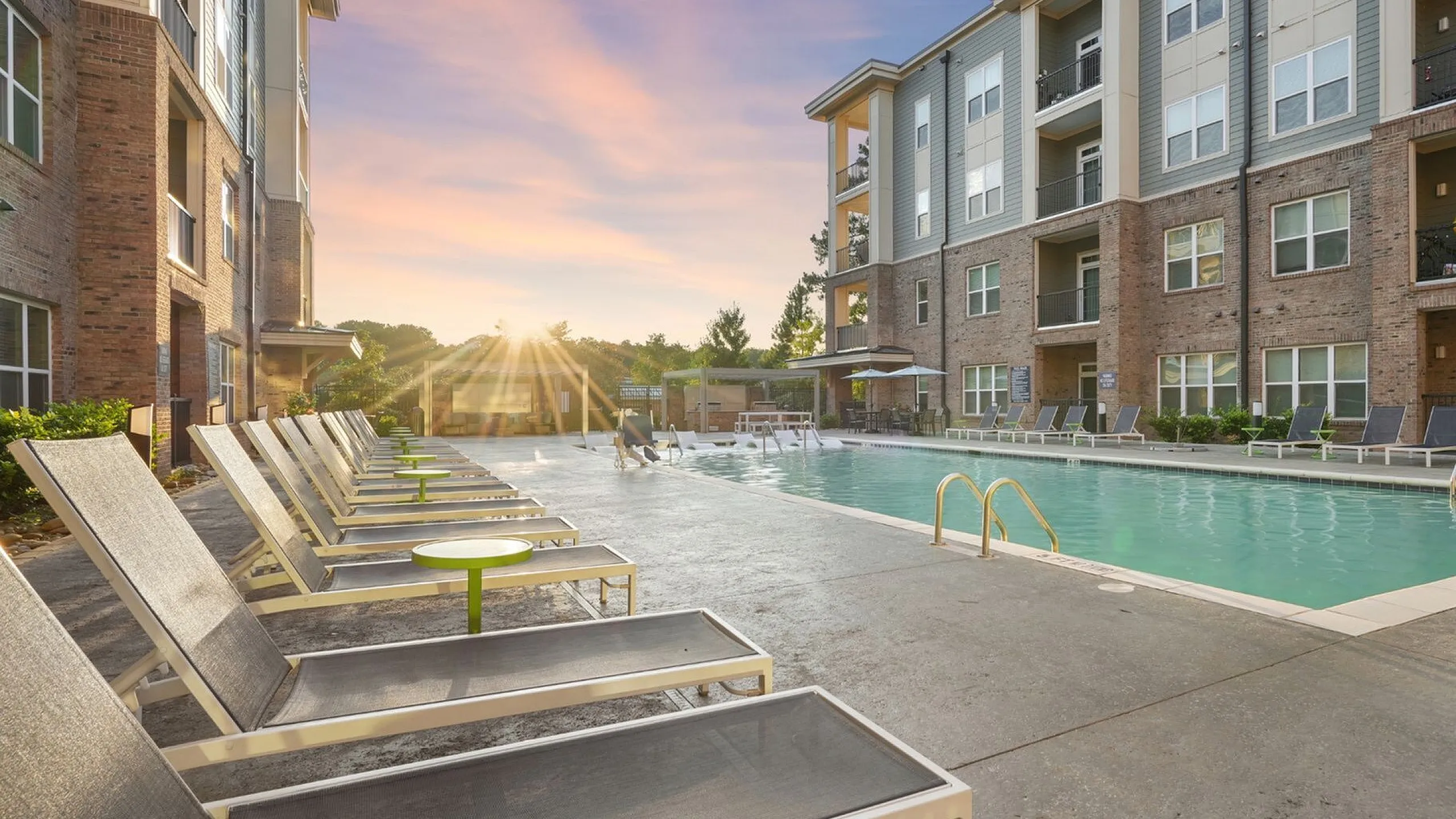 Inviting apartment complex with a luxurious swimming pool, surrounded by modern lounge chairs and a stunning sunset view.