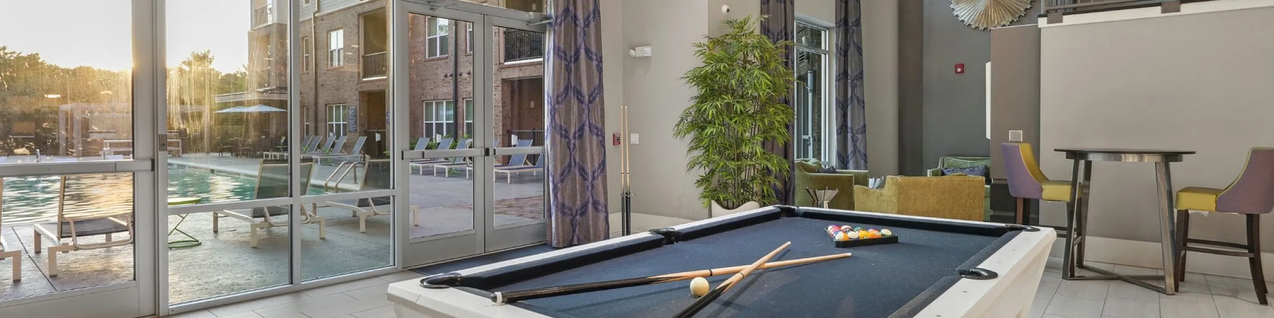Luxurious recreation area with a stylish pool table, high ceilings, and large windows overlooking a beautiful pool. Sophisticated decor includes modern wall art, greenery, and chic seating for relaxation.
