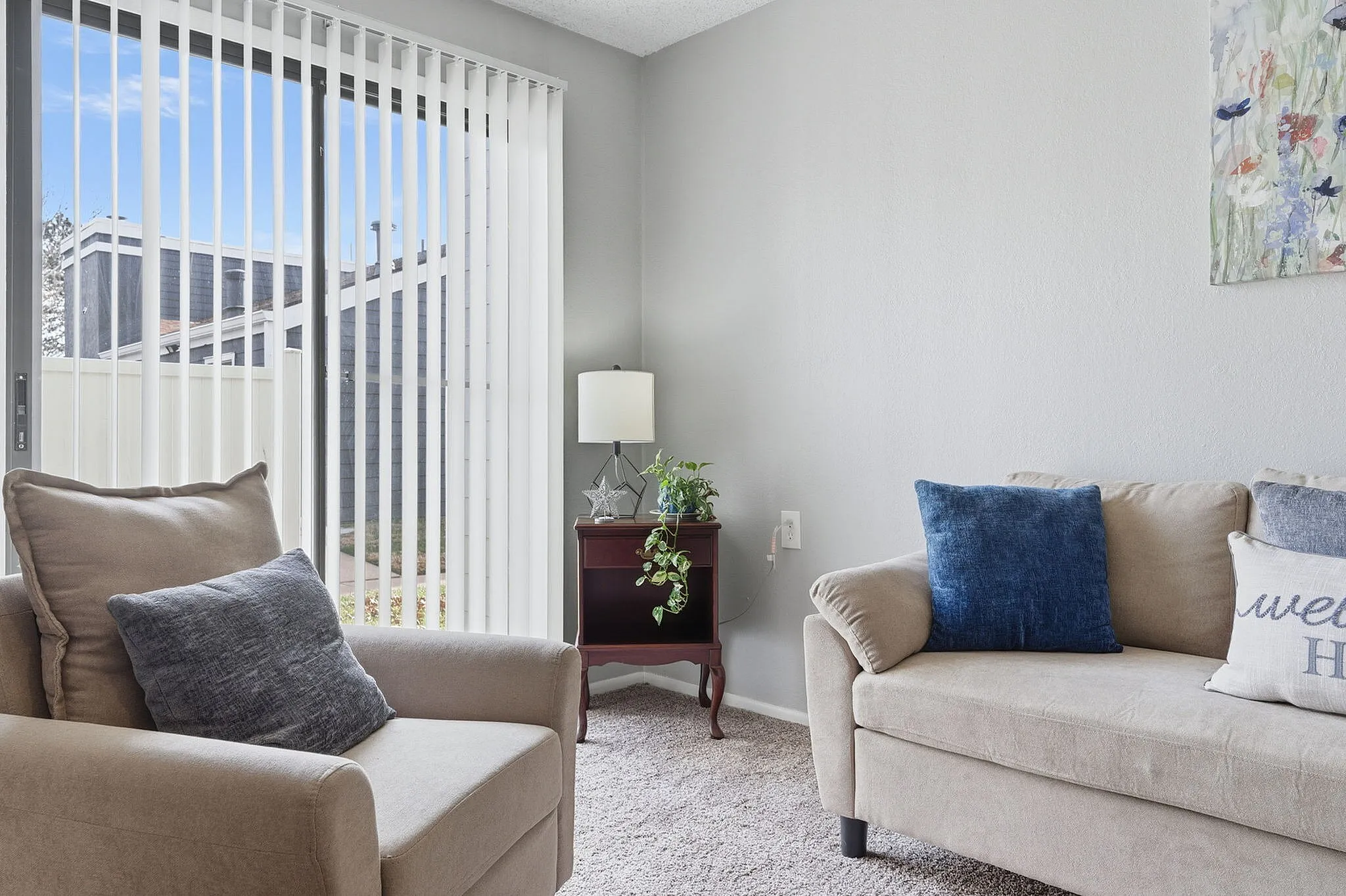 Bright and inviting living room with cozy beige sofas, vibrant blue cushions, and a sunlit view through large vertical blinds.