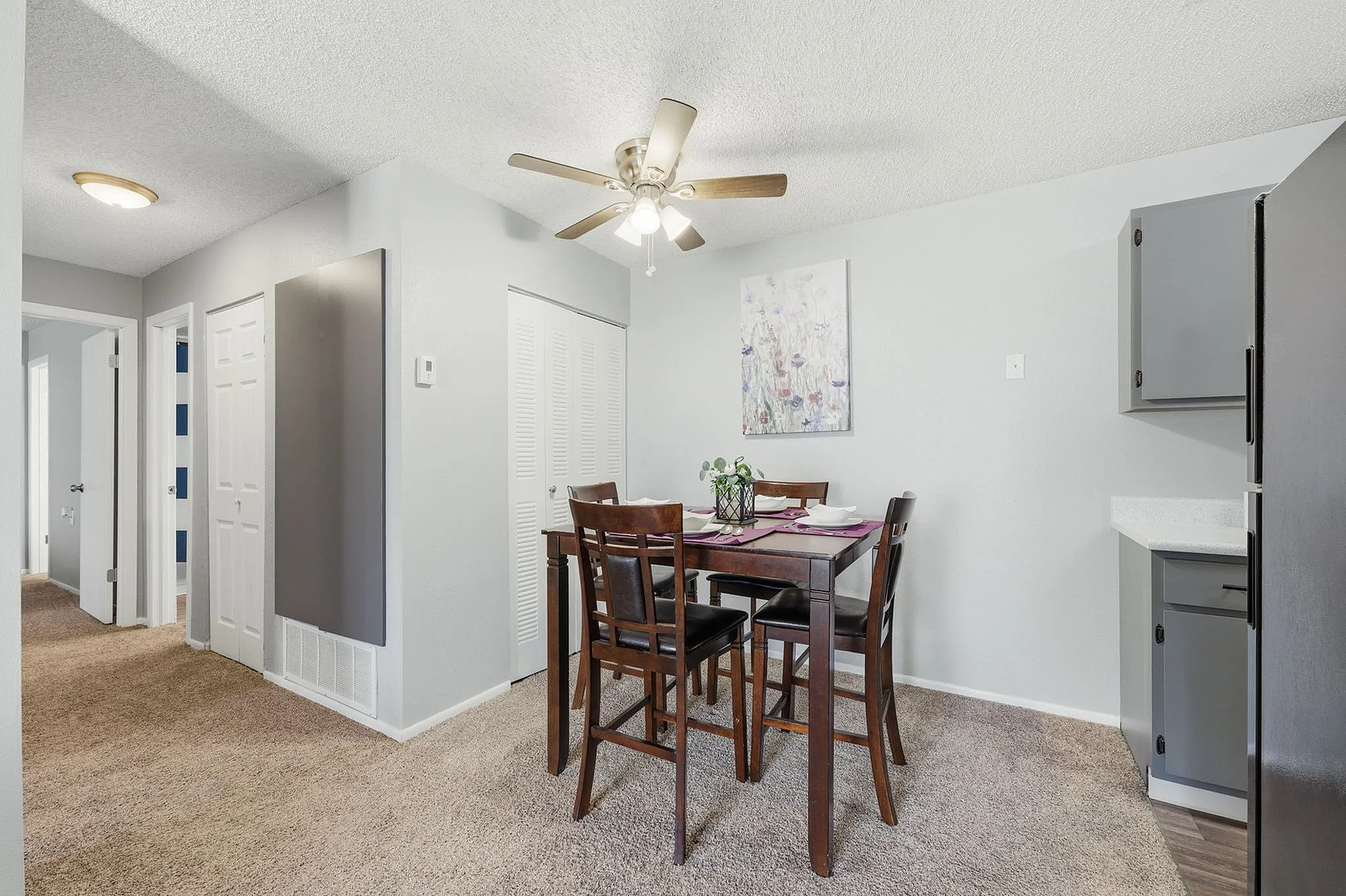 Modern dining area with a stylish table set under a ceiling fan, surrounded by soft carpet and contemporary art, leading into a bright hallway.