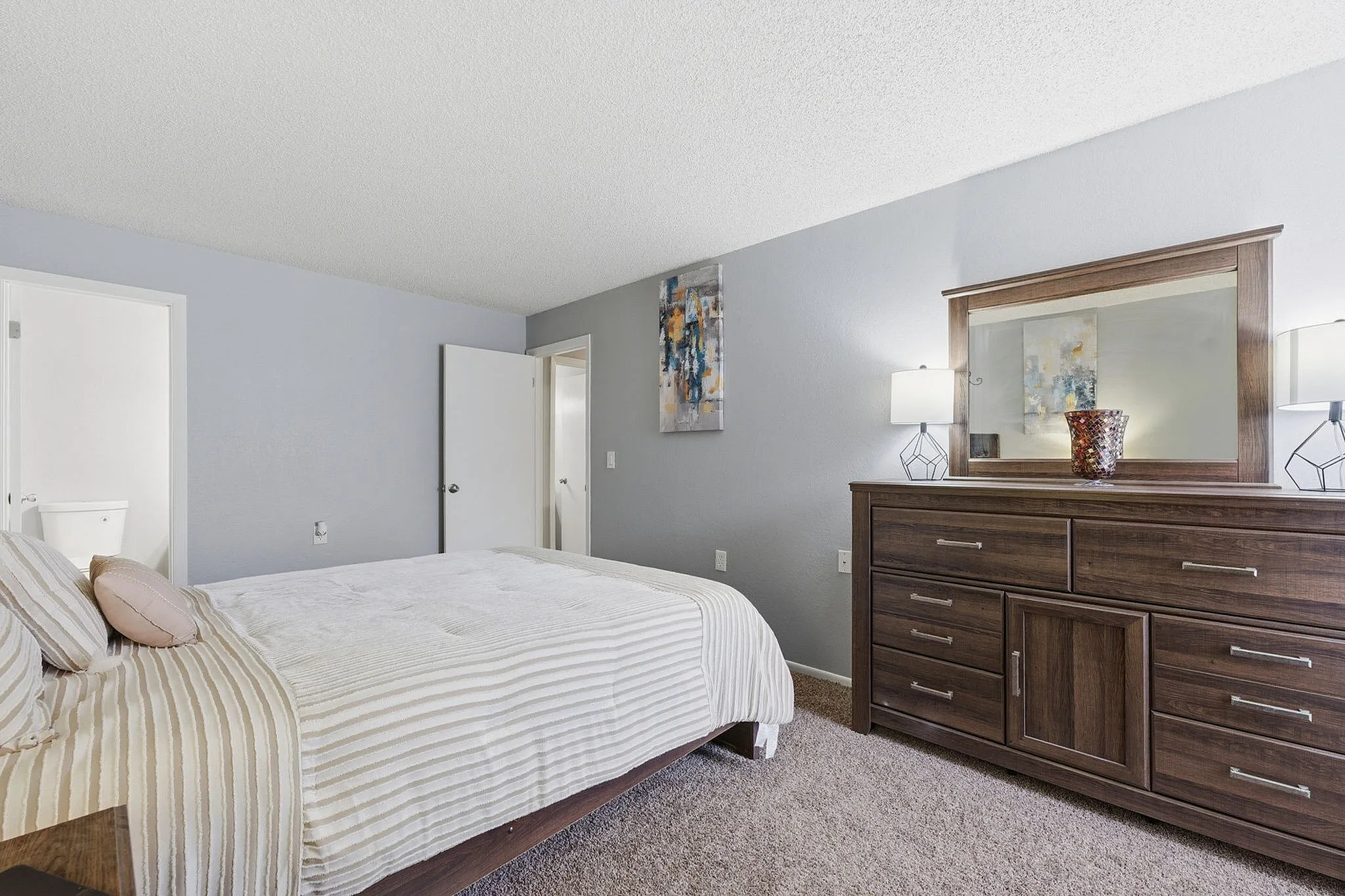 Inviting bedroom with plush carpeting, elegant dresser with mirror, and cozy bed adorned with striped bedding, creating a warm and tranquil retreat.
