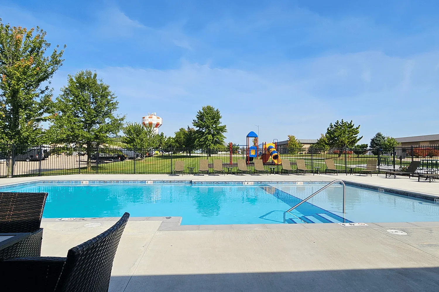 Inviting outdoor pool area with sun loungers, adjacent to a colorful children's playground under a clear blue sky.