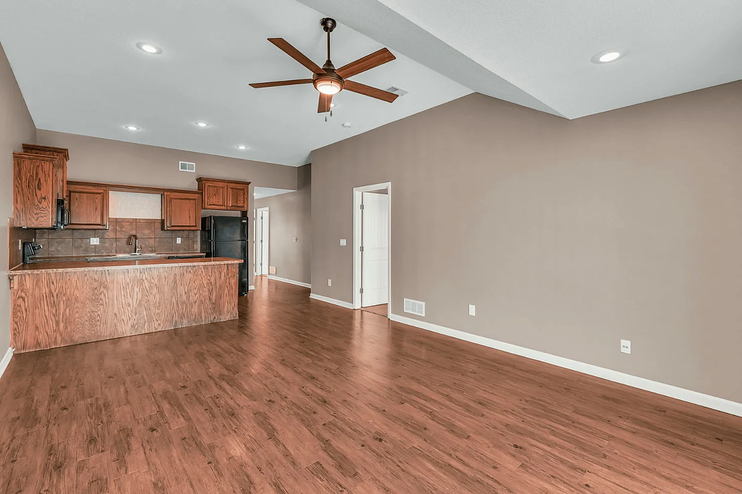 Spacious modern apartment living area with warm wooden flooring and sleek ceiling fan, adjacent to a stylish open kitchen with elegant wood cabinets.