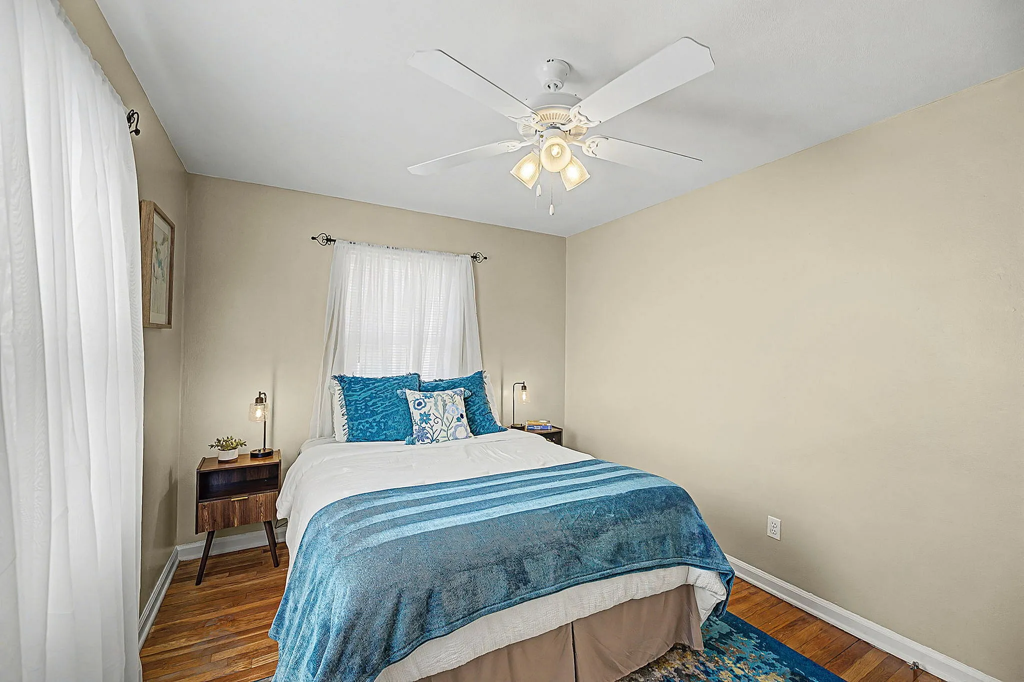 Inviting bedroom with elegant bedding, wooden floor, a stylish ceiling fan, and soothing natural light.