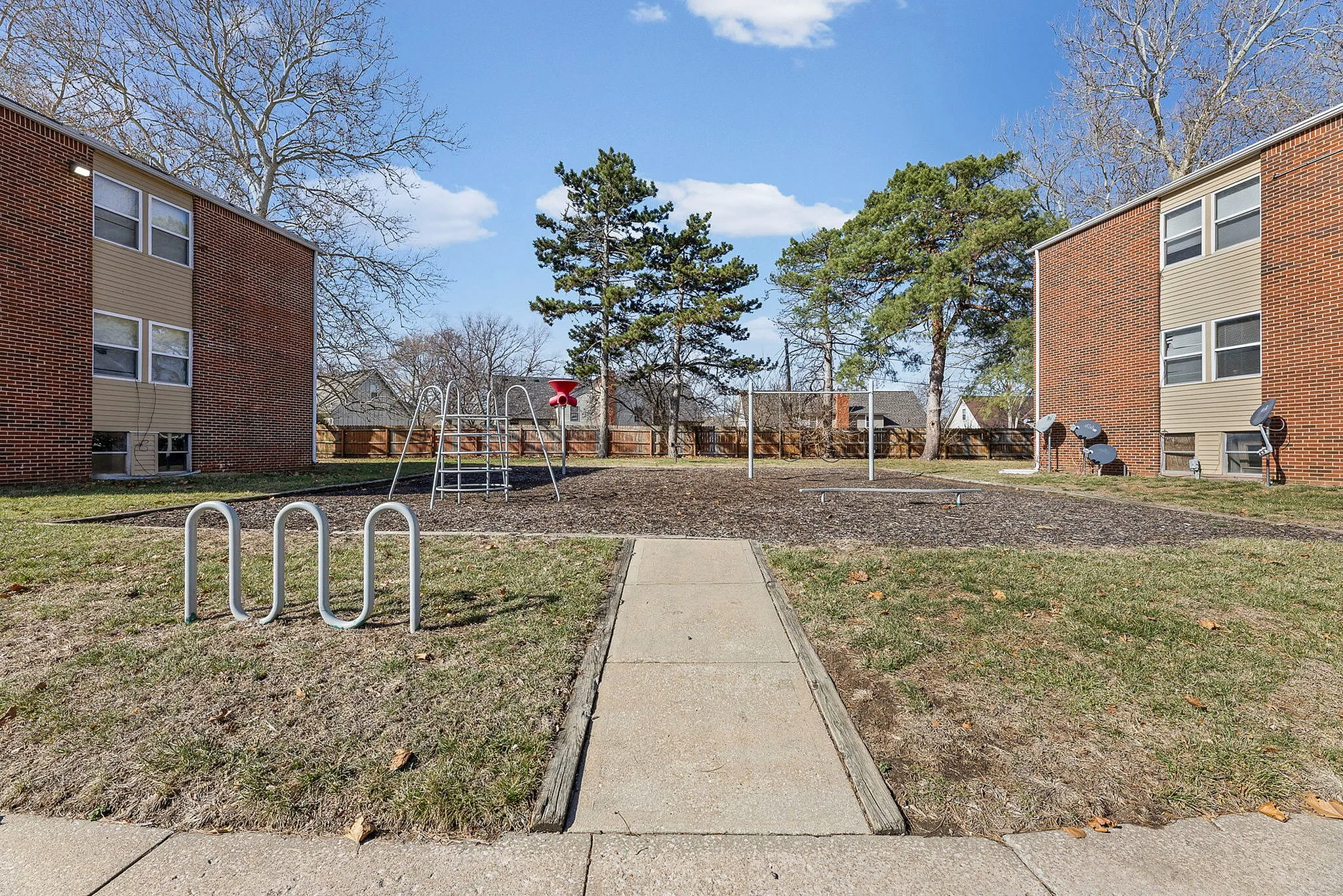 Charming outdoor playground area nestled between brick apartment buildings with lush trees and clear blue skies.