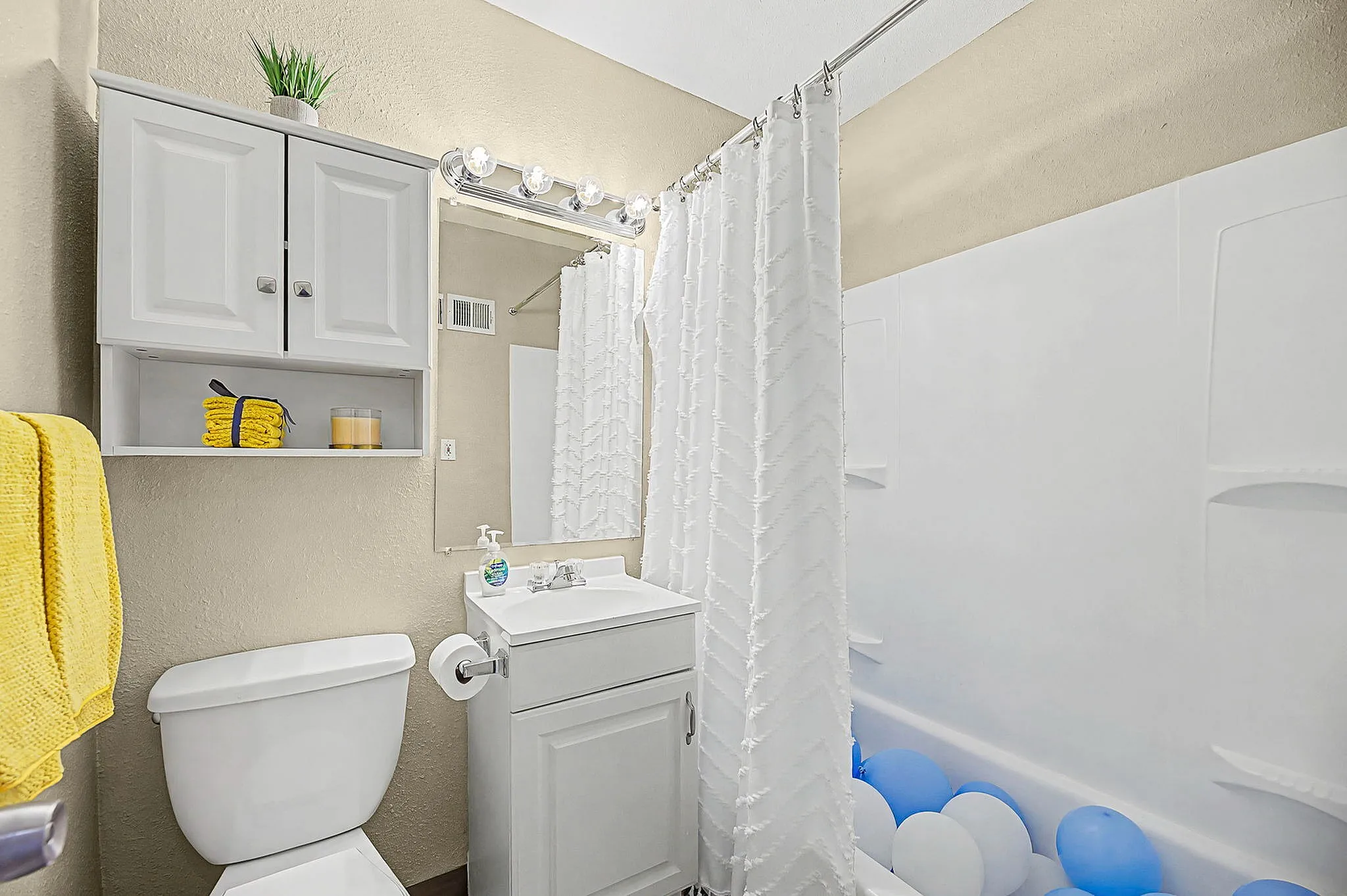 Bright and inviting bathroom featuring modern white cabinets, a sleek vanity with storage, vibrant yellow towels, and a playful touch of blue and white balloons in the tub, creating a cheerful ambiance.