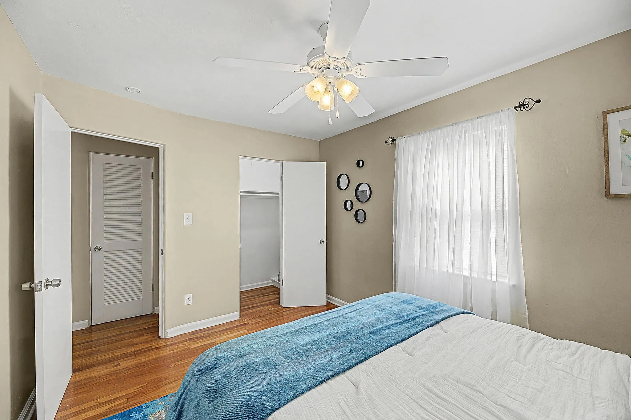 Inviting bedroom with warm wooden floors, a cozy bed adorned with a blue throw, and soft natural light filtering through sheer curtains.