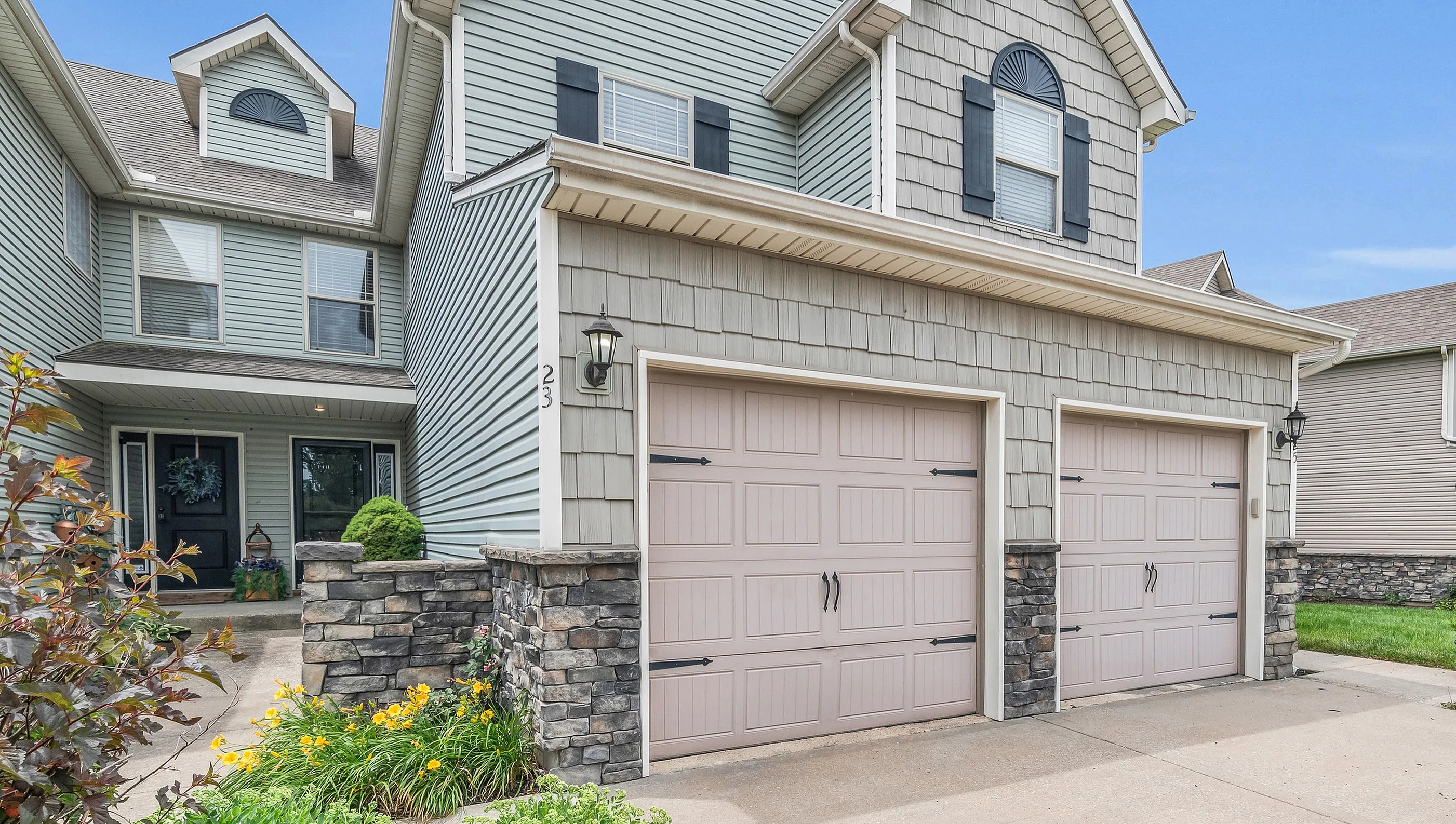 Charming suburban home with dual beige garage doors, elegant stone accents, and vibrant landscaping.