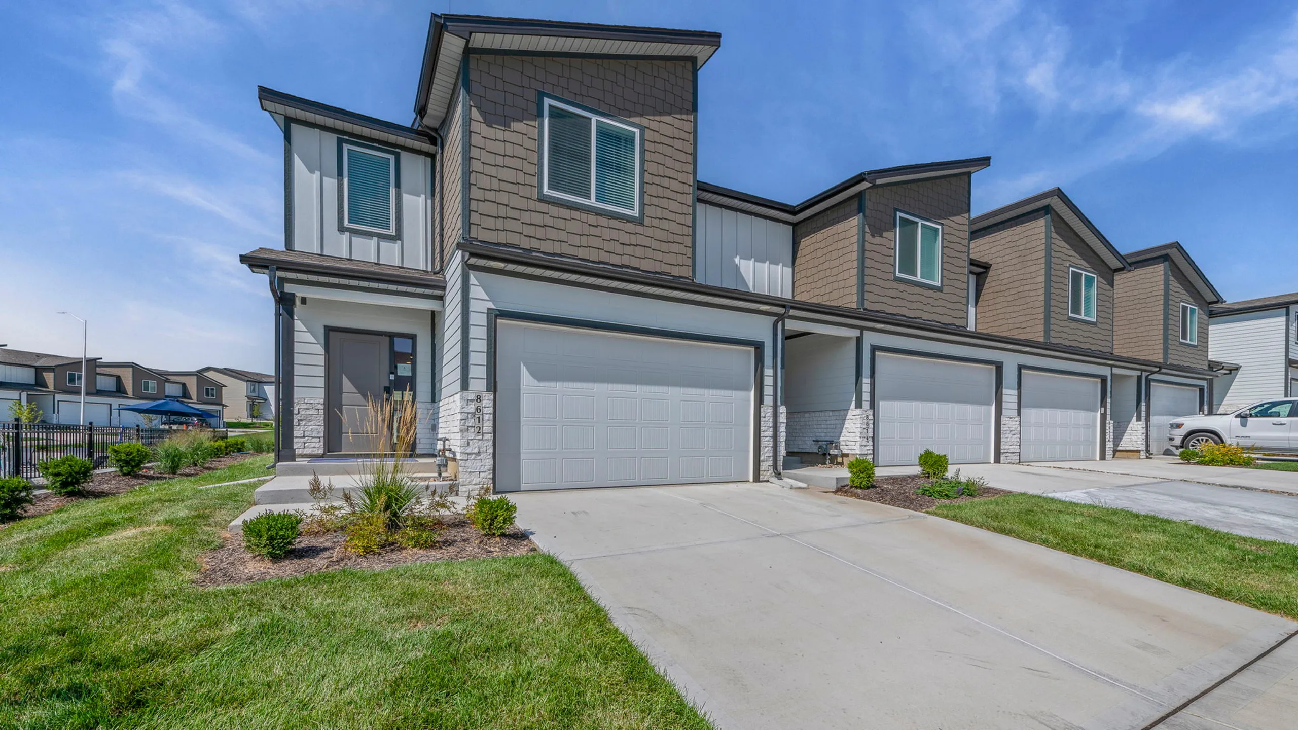 Charming townhouse exterior with modern design, featuring a two-story layout and attached garage, surrounded by lush landscaping and clear blue skies.