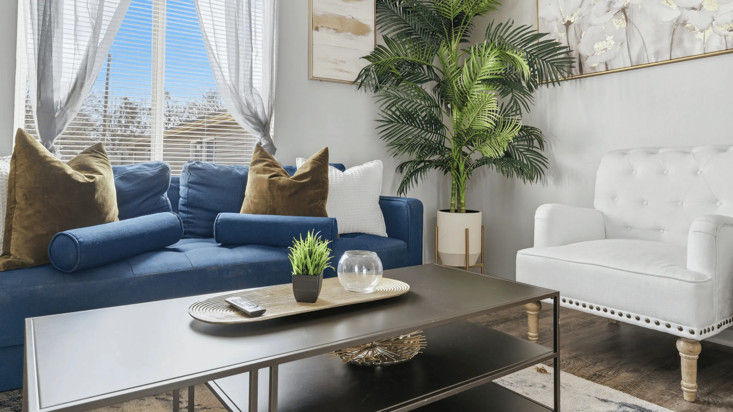 Bright and stylish living room with a plush blue sofa, chic white armchair, and vibrant green plant, complemented by elegant decor and large windows allowing abundant natural light.