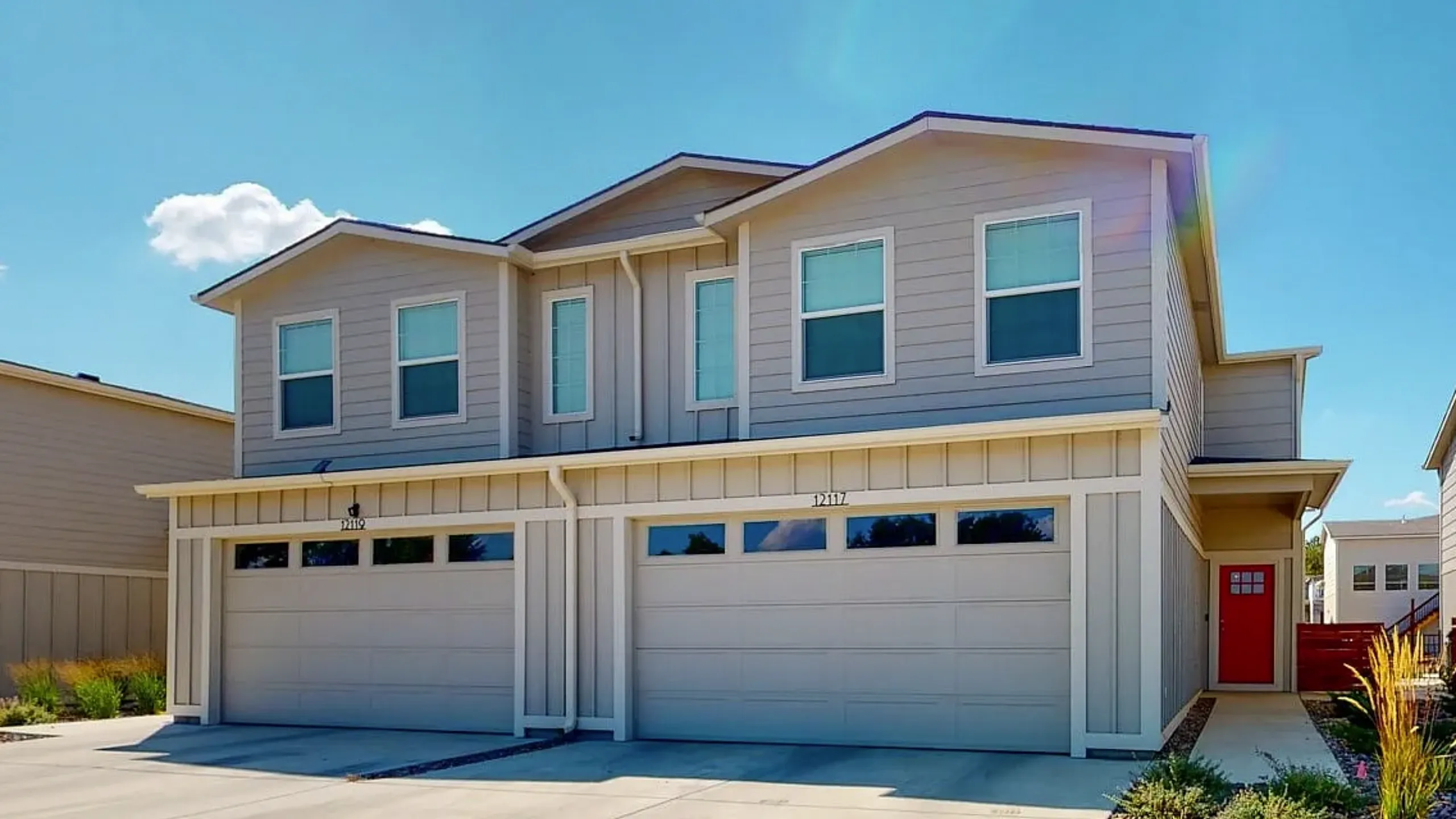 Modern duplex with sleek design, featuring dual garages and vibrant red front door under a bright sky.