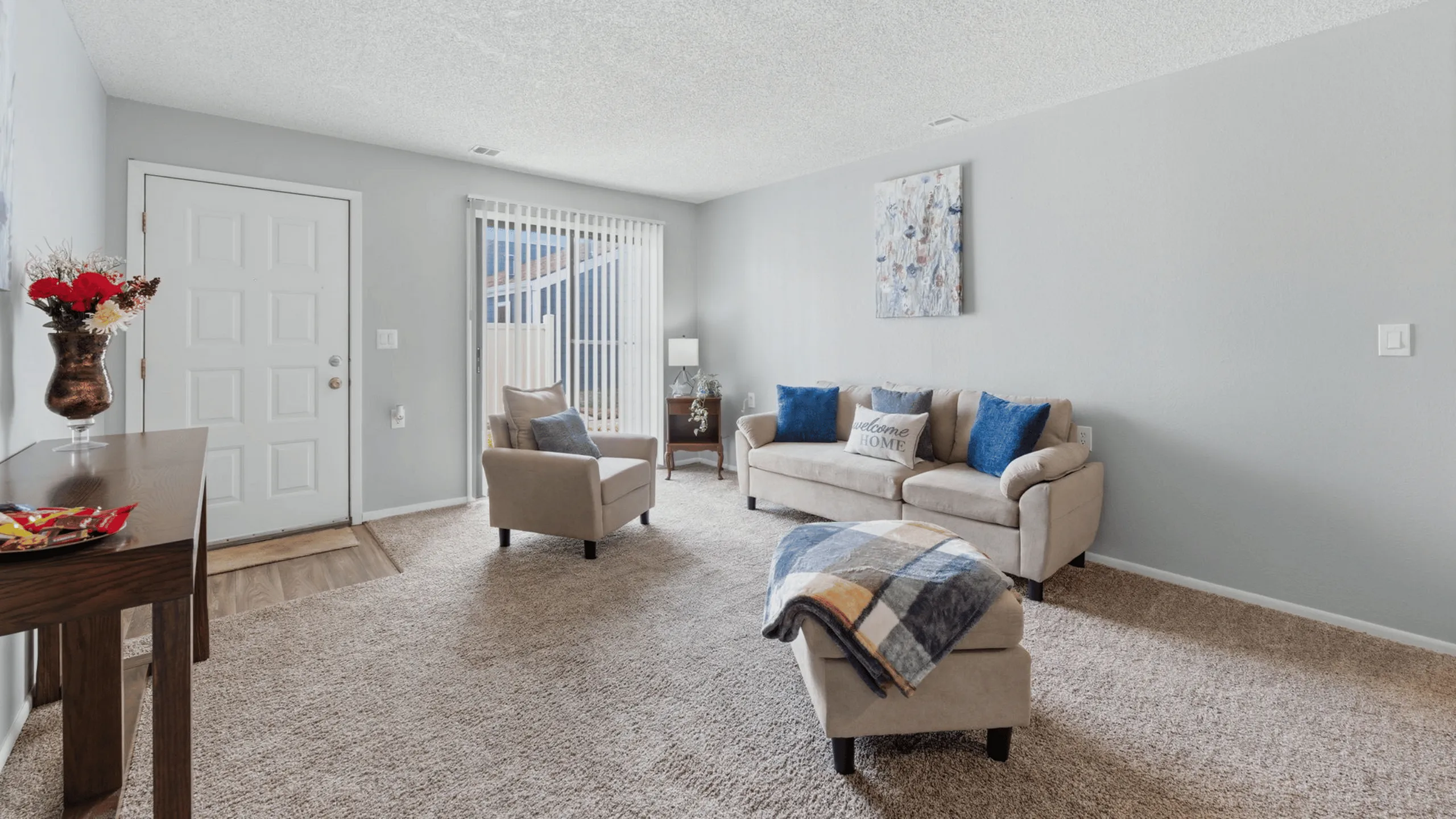 Inviting living room with cozy beige sofas, vibrant blue cushions, and elegant decor, accented by a welcoming entrance with natural light streaming through vertical blinds.