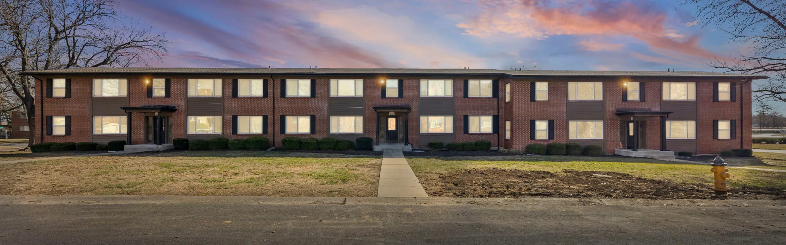 Charming brick apartment building with modern black shutters, set against a vibrant sunset sky. Ideal for comfortable living.