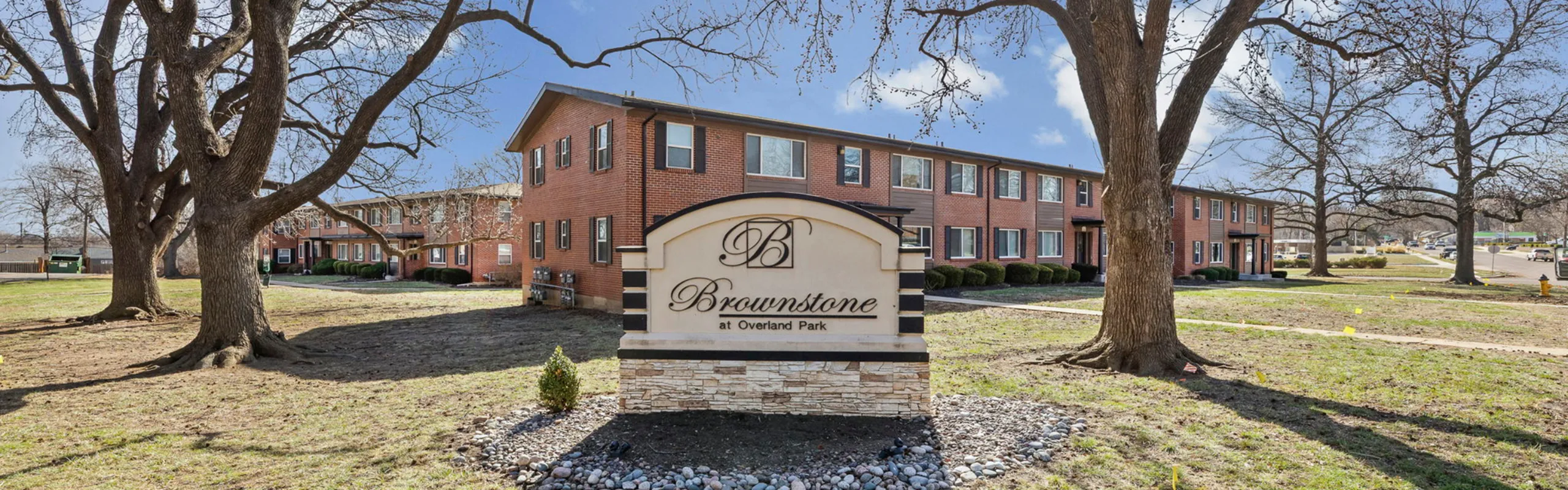 Charming brick apartment complex with a welcoming landscaped entrance at Brownstone, surrounded by mature trees in a peaceful neighborhood.
