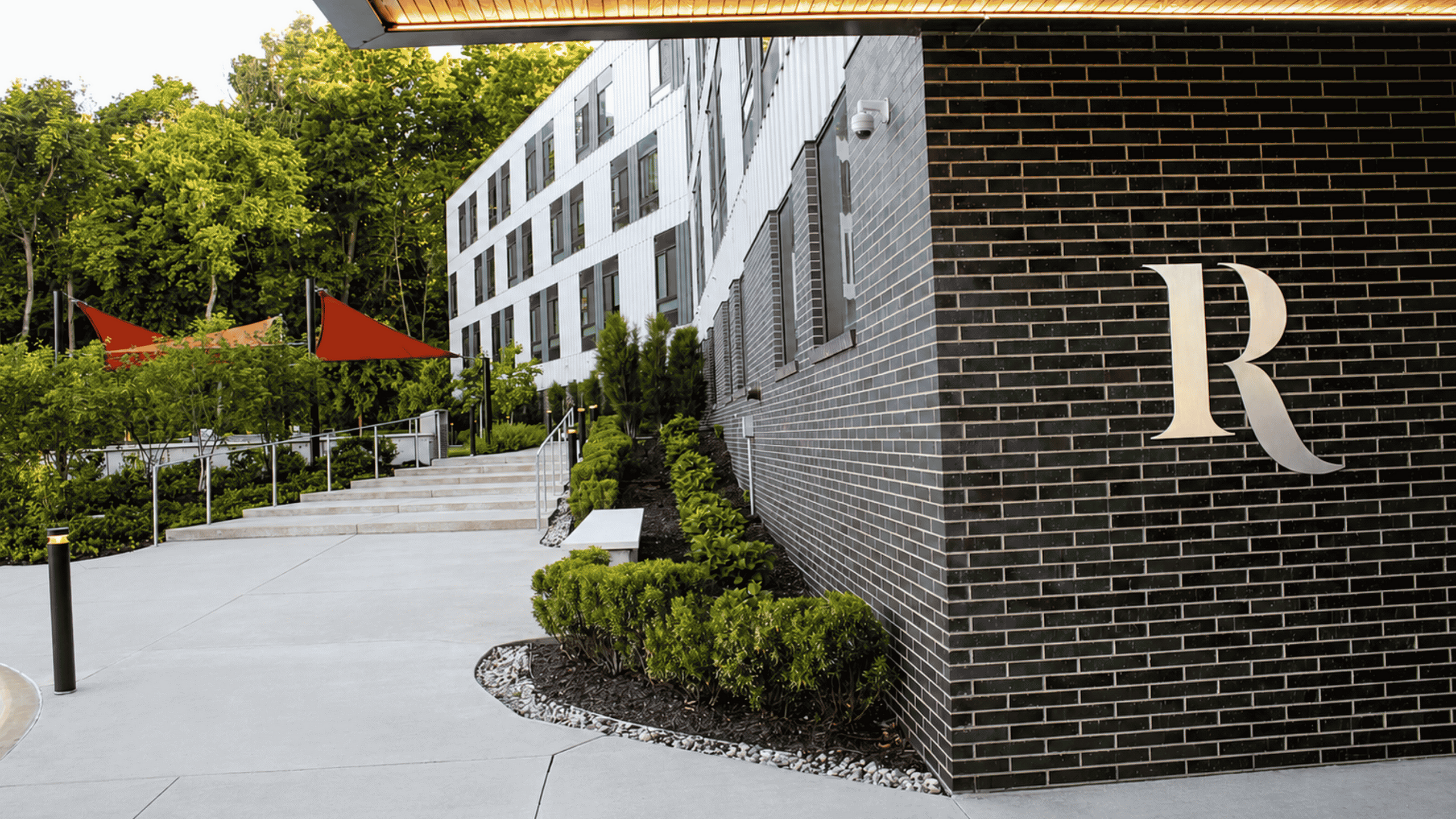 Chic modern apartment exterior with stylish brick facade and lush landscaping, offering a serene entrance with vibrant red shade sails and welcoming pathway.