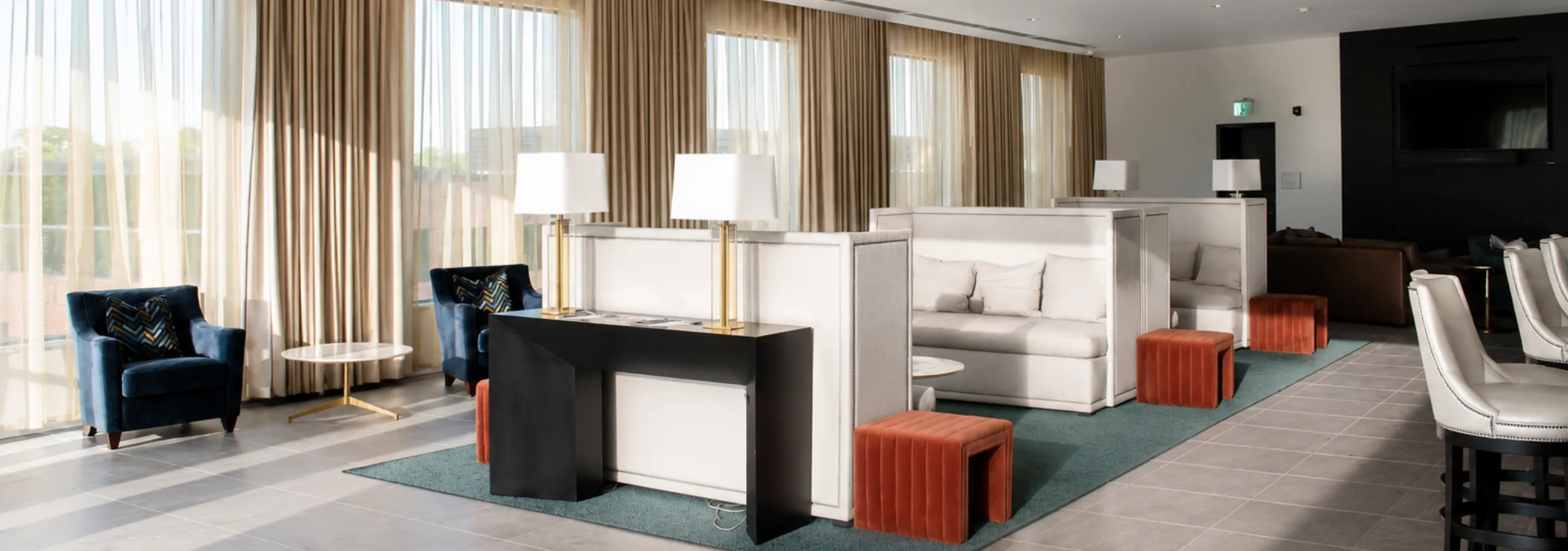 Elegant lounge area featuring modern sofas, vibrant orange stools, plush blue armchairs, and chic white bar stools, bathed in natural light from floor-to-ceiling windows with flowing curtains.
