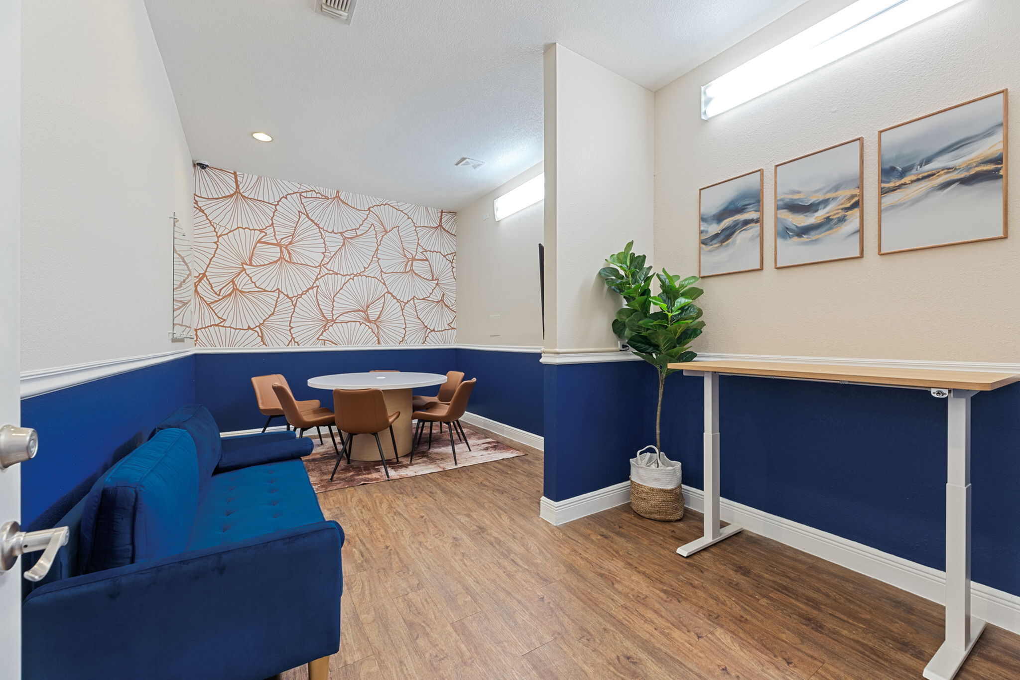 Chic room interior with a stylish blue sofa, modern round table with tan chairs, artistic wallpaper, and elegant wall art, complemented by vibrant green plants.