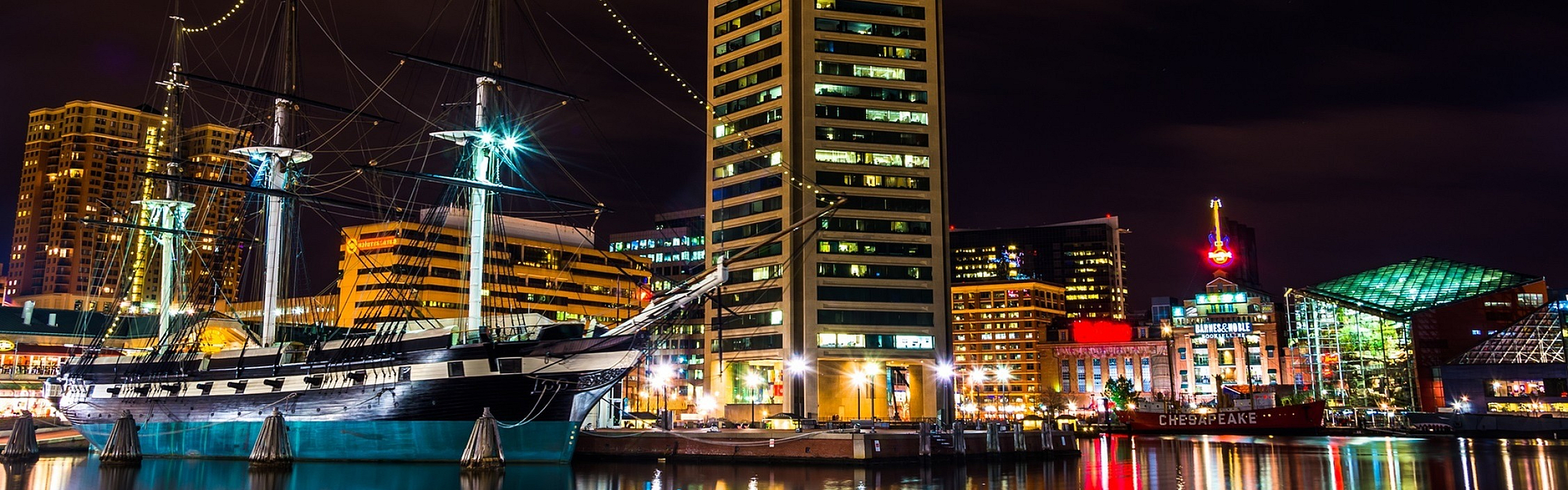 Stunning waterfront night view featuring a historic ship next to a modern high-rise, illuminated cityscape reflecting on calm waters.