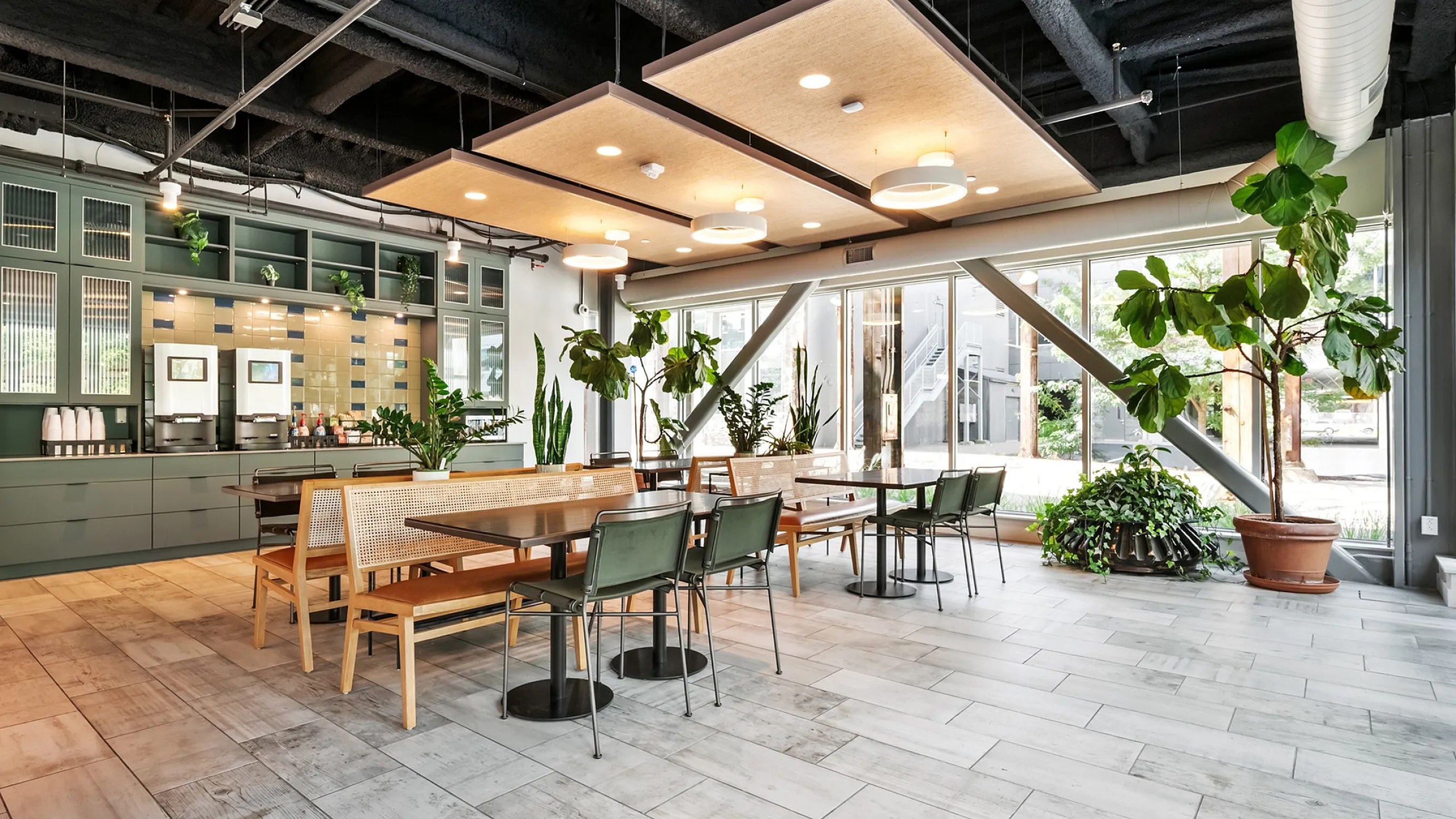 Modern communal space with sleek tables, lush greenery, and a contemporary coffee station under chic ceiling panels.