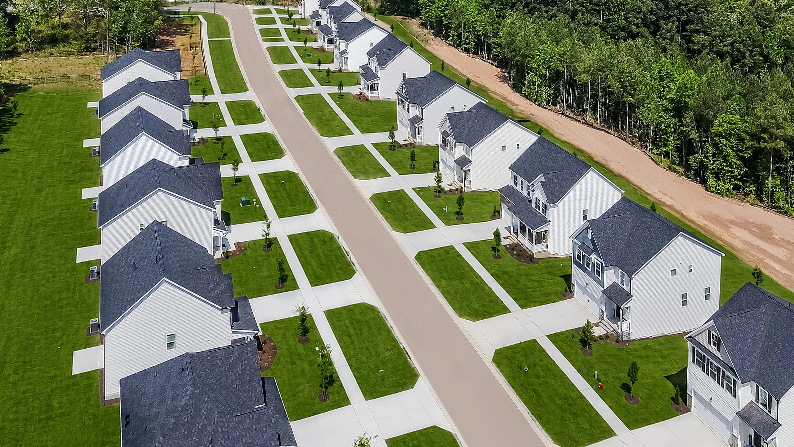 Scenic aerial view of a modern suburban neighborhood with rows of charming, white-painted homes, lush green lawns, and a backdrop of tranquil forested areas. Ideal living space for families seeking serenity.