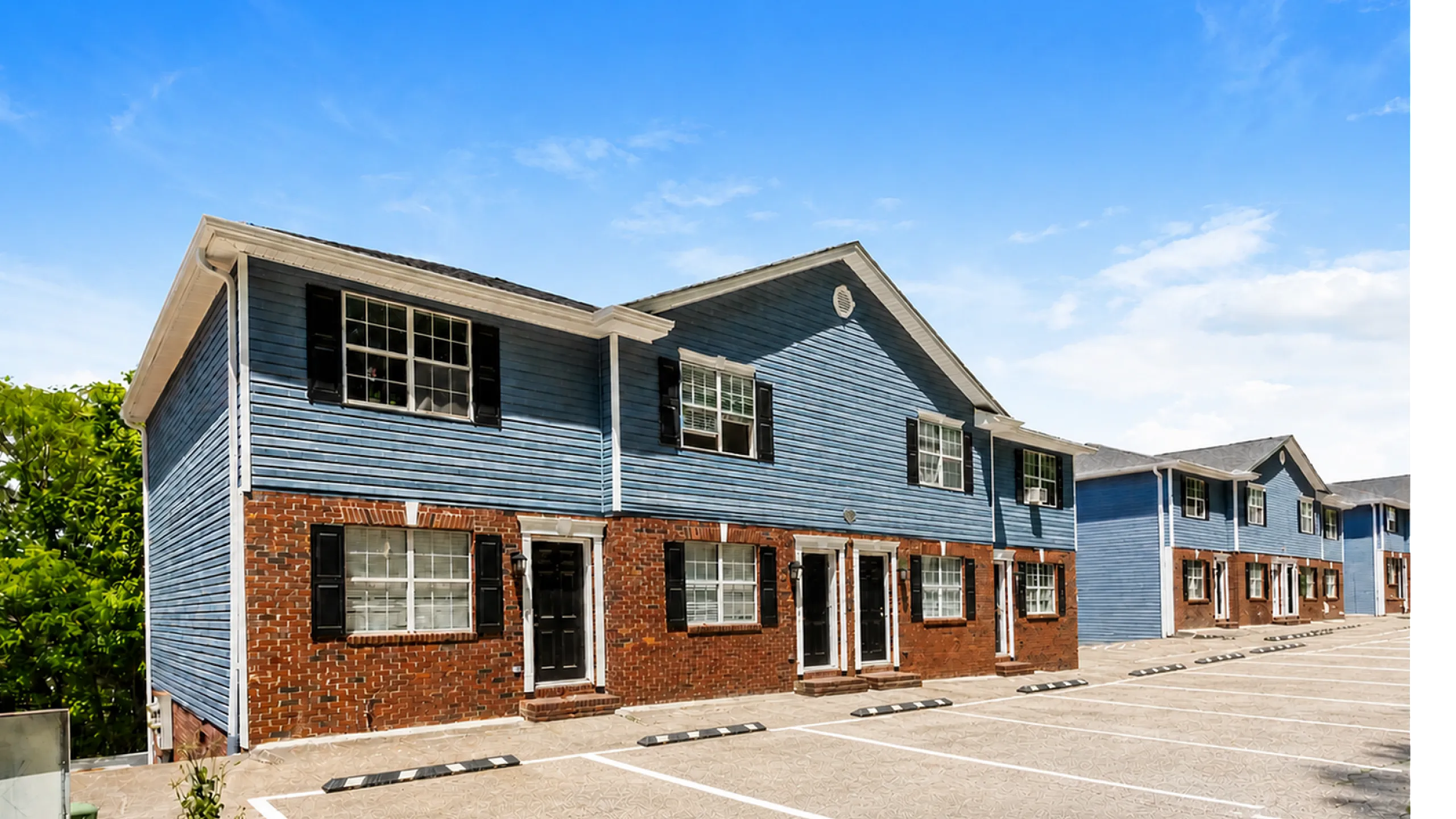 Charming townhouse complex with blue siding and brick accents, featuring ample parking and lush greenery under a clear blue sky.