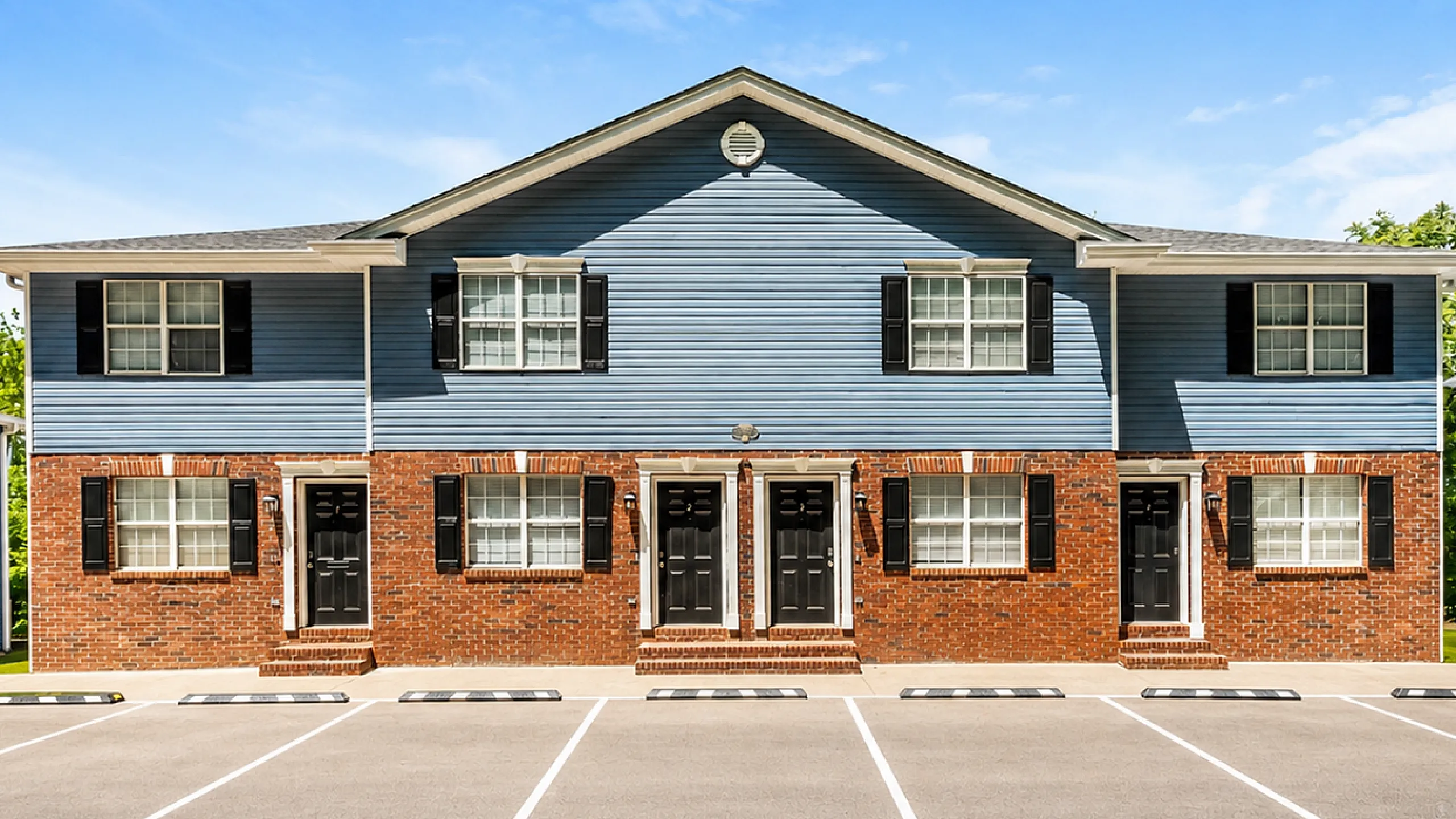 Charming blue multi-unit apartment building with brick accents and ample parking space, set under a clear blue sky.