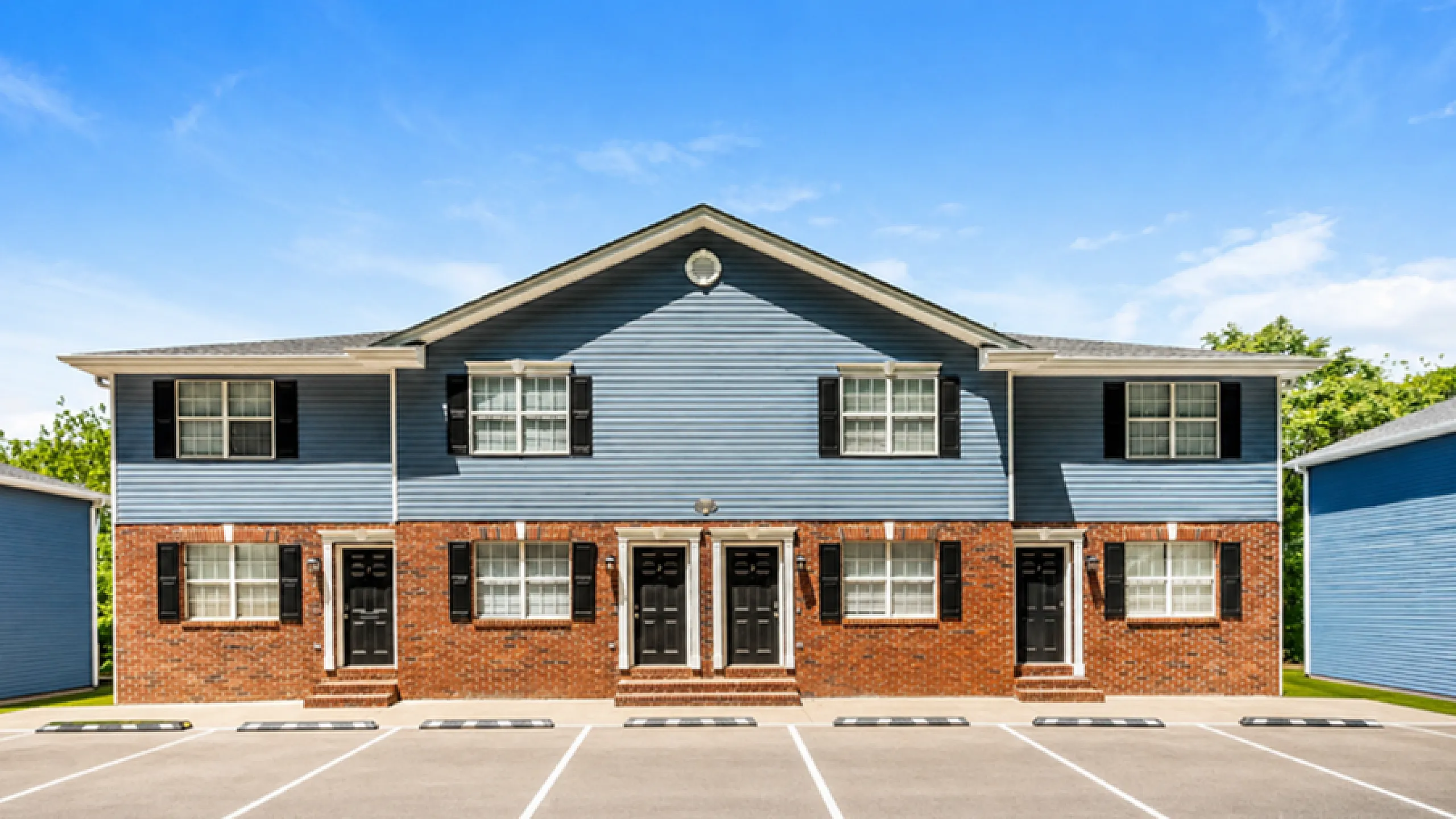 Charming townhouse exterior with classic brick facade and blue siding, offering ample parking and lush surroundings under a clear blue sky.