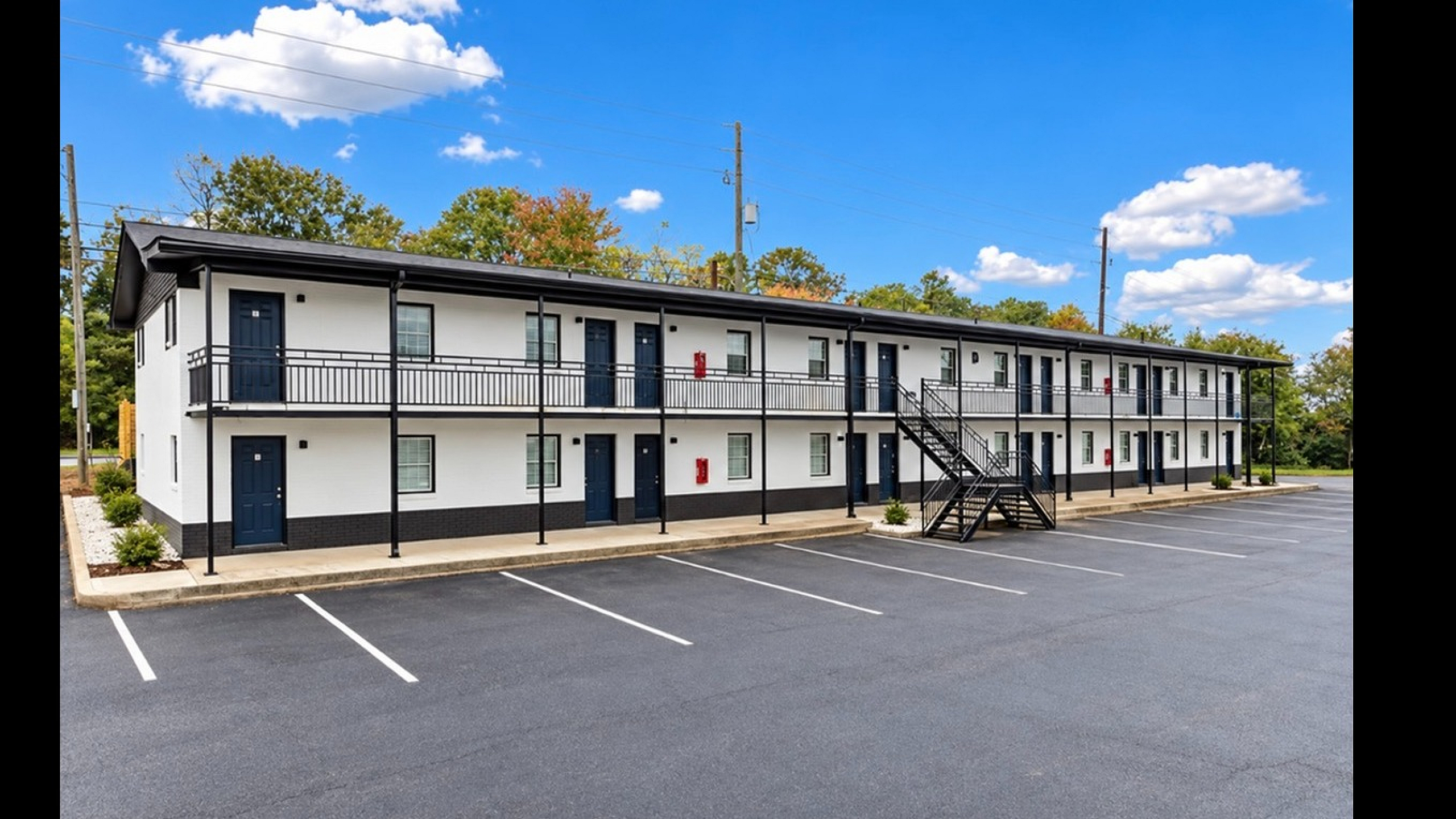 Modern two-story apartment complex with fresh white and black exterior, featuring ample parking space and surrounded by lush greenery under a clear blue sky.