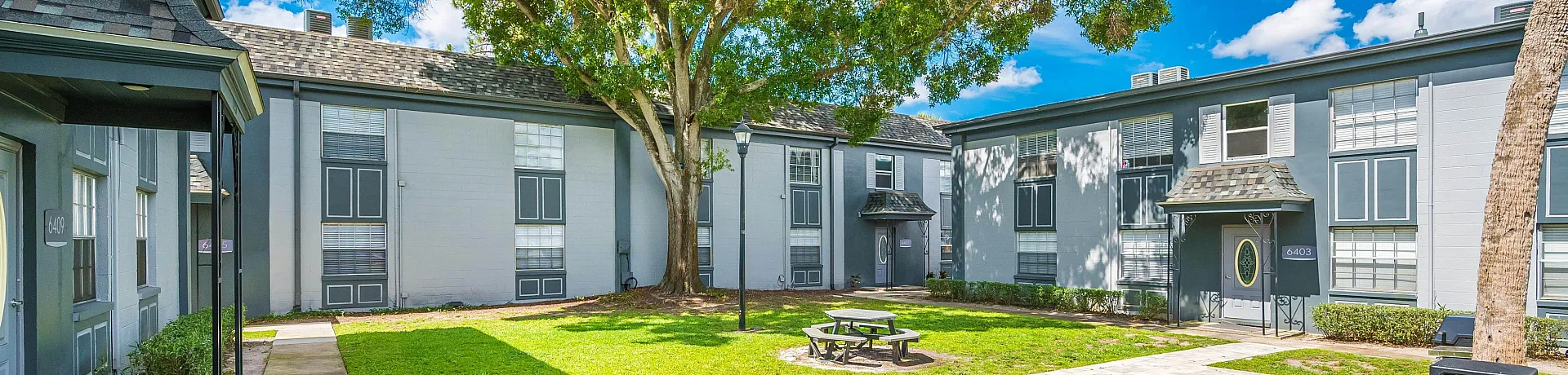 Charming apartment courtyard with lush greenery, picnic table, and spacious walkways under a bright blue sky.