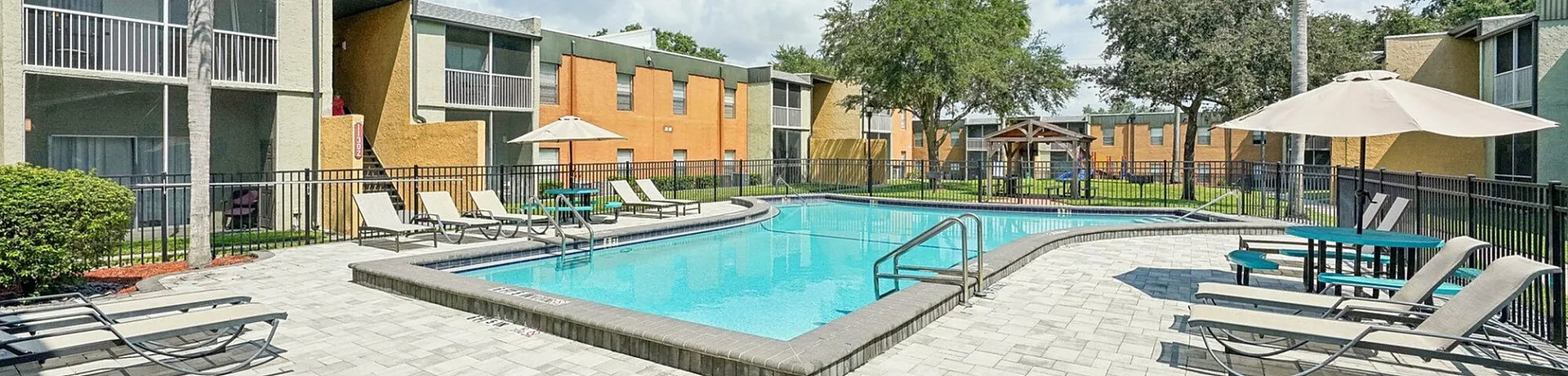 Inviting apartment complex with a sparkling outdoor pool, surrounded by stylish lounge chairs and lush palm trees under a vibrant blue sky.