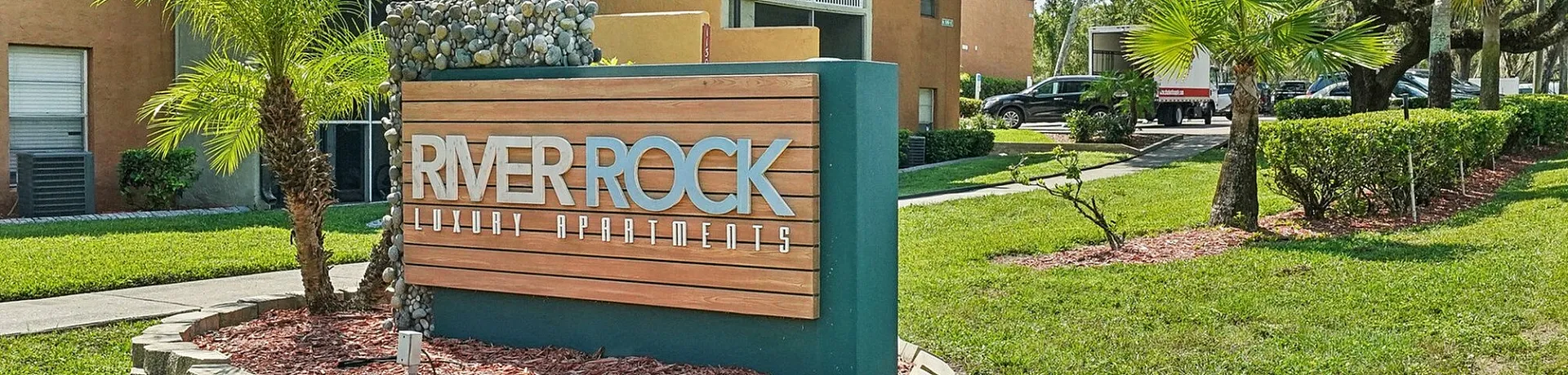 Charming exterior view of River Rock Luxury Apartments with lush greenery and attractive landscaping, showcasing a welcoming entrance sign.