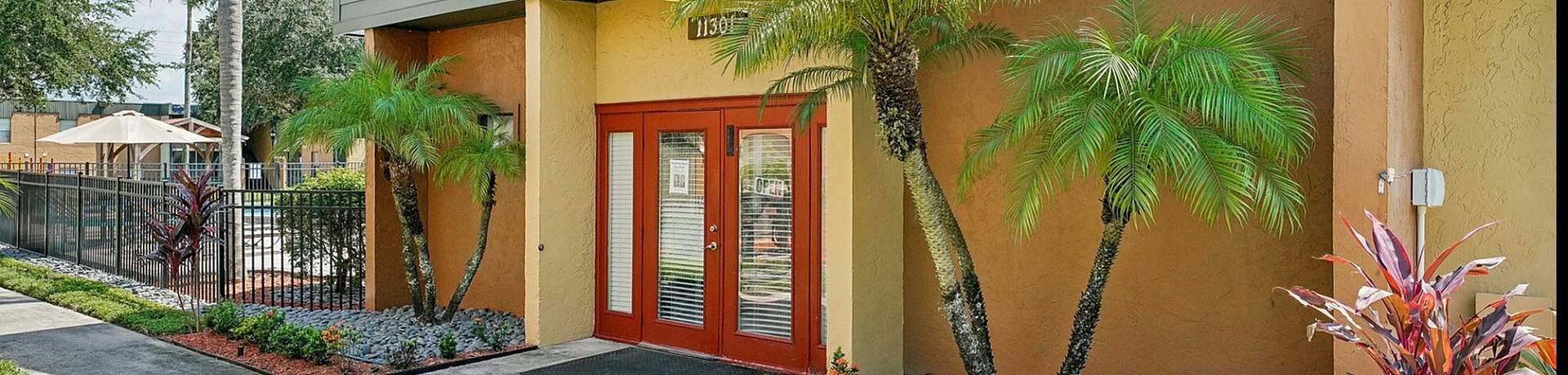 Charming apartment entrance with vibrant landscaping, tropical plants, and inviting red doors under a clear blue sky.