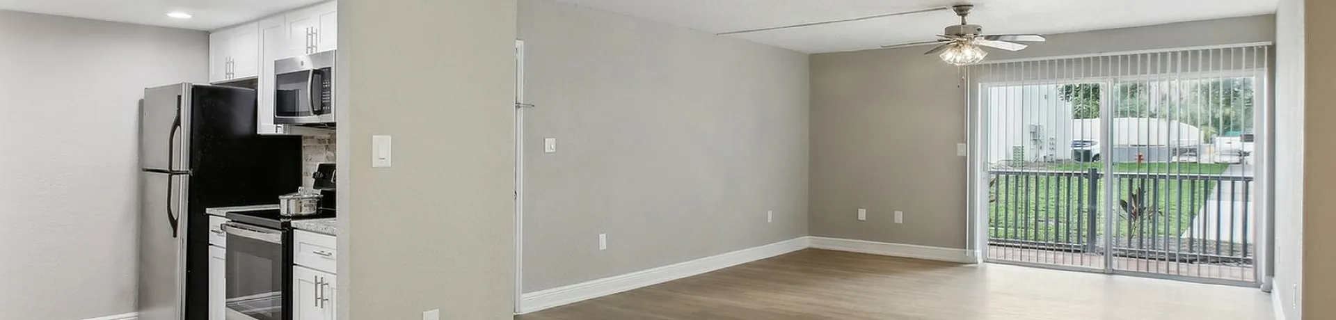 Bright and spacious living area with sleek wood flooring, modern kitchen appliances, and large sliding glass doors opening to a lush green view.