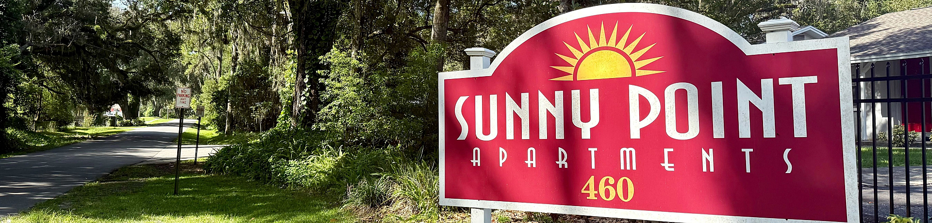 Welcome to Sunny Point Apartments, nestled in a lush green setting, offering a serene and inviting atmosphere with a charming entrance sign.