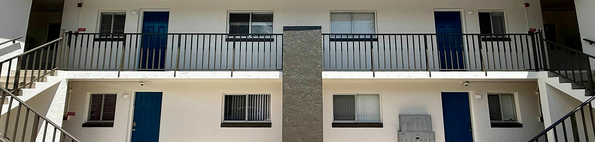 Charming two-story apartment building with blue doors and a welcoming courtyard, surrounded by clear skies.