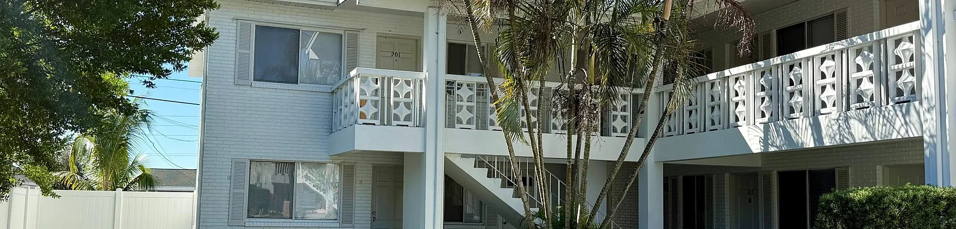Charming apartment building with unique balcony railings, surrounded by lush greenery and a barbecue area on a sunny day.