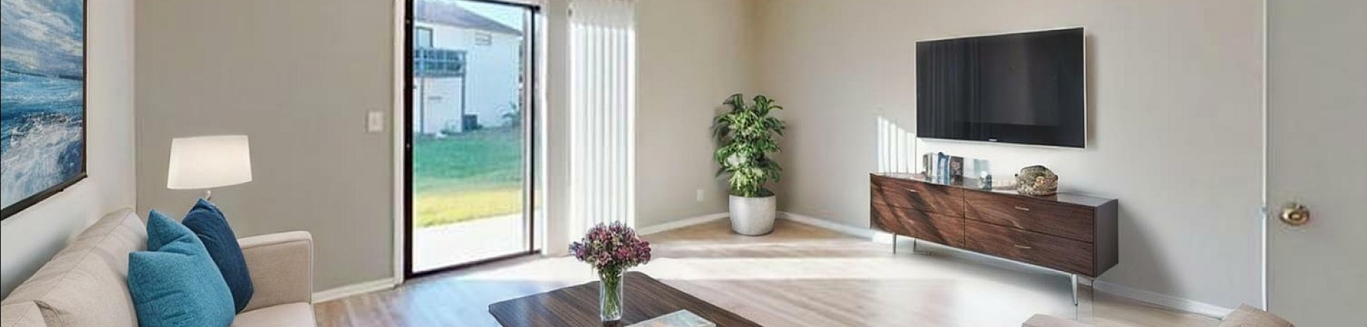 Bright living room with sliding glass doors leading to a lush backyard, featuring a cozy sofa with blue cushions, stylish coffee table, and wall-mounted TV, perfect for relaxation and entertaining.