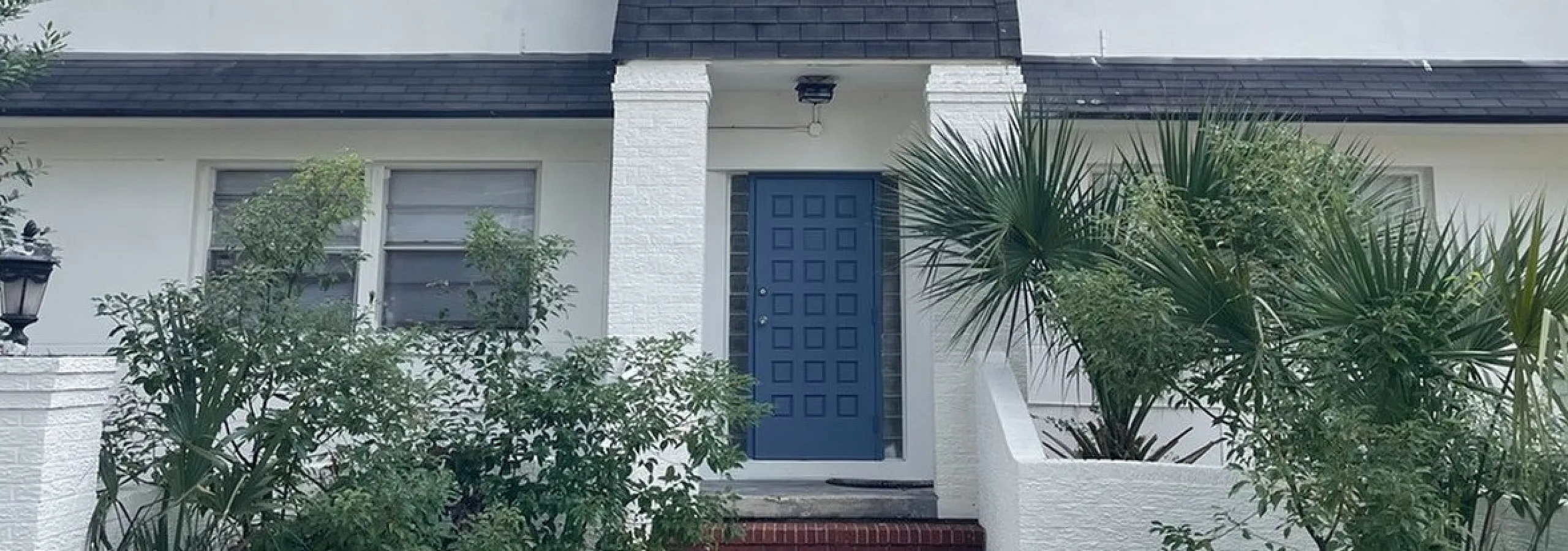 Charming two-story white house with a classic blue door, surrounded by lush greenery and a welcoming pathway.