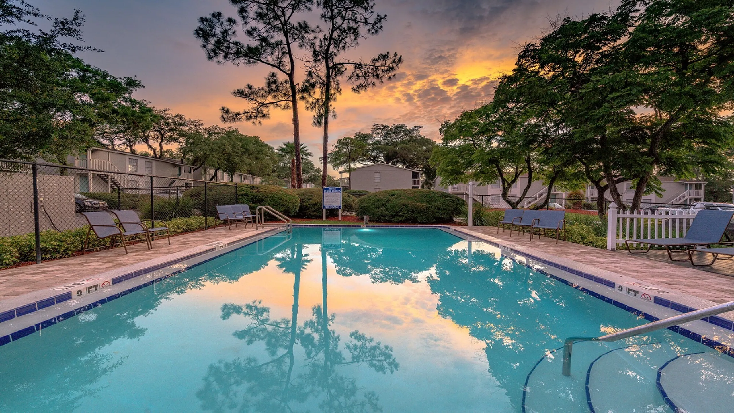 Serene apartment community pool surrounded by lush greenery and stylish lounge chairs, reflecting a vibrant sunset sky.