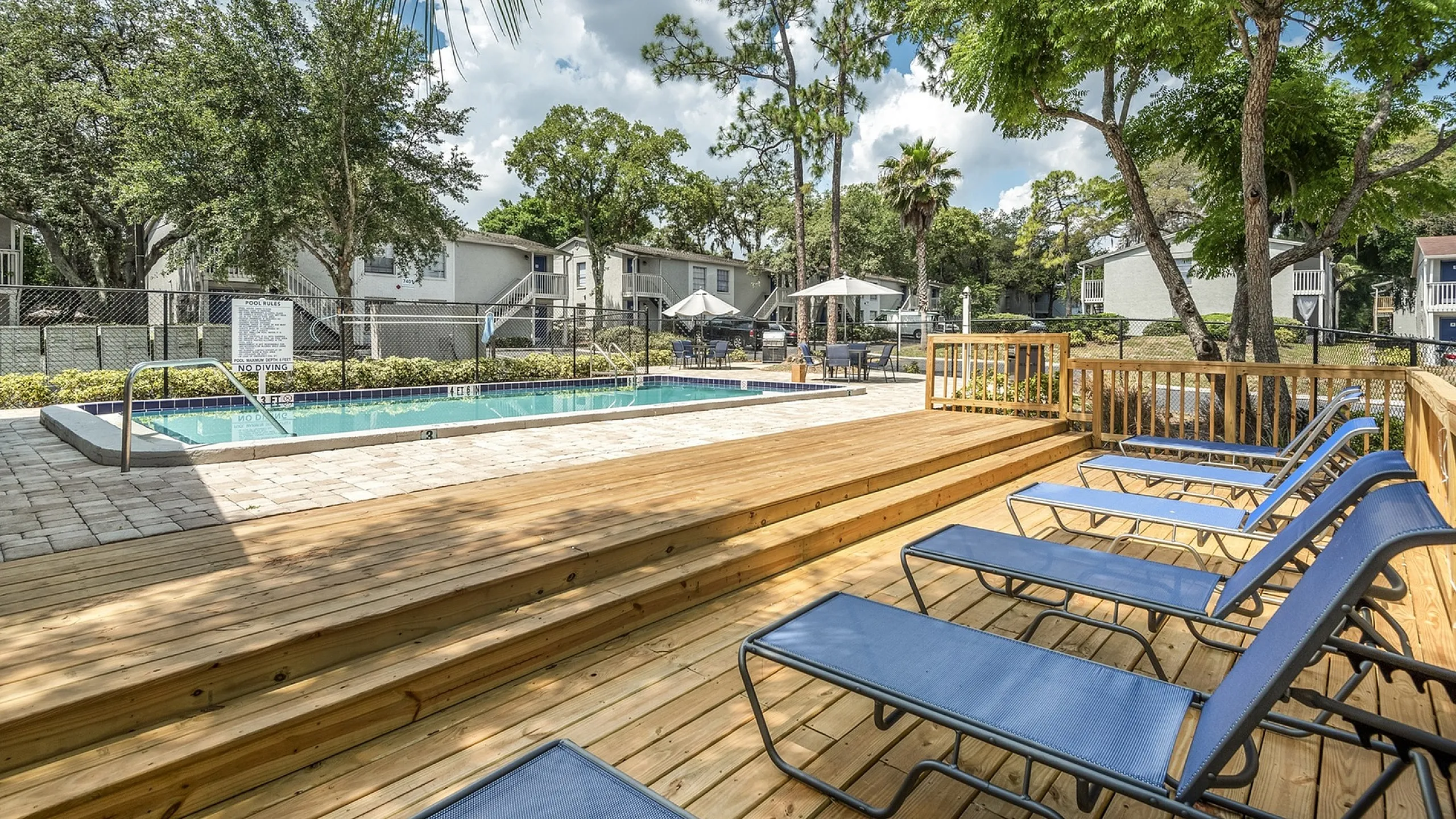 Charming outdoor pool area with wooden deck, comfortable loungers, and lush green surroundings in a serene residential community.