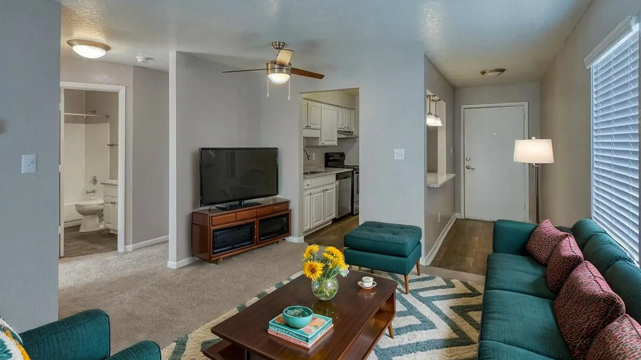 Inviting living room with modern decor, featuring a comfortable teal sofa, stylish wooden coffee table, and a flat-screen TV beside an elegant kitchen area.